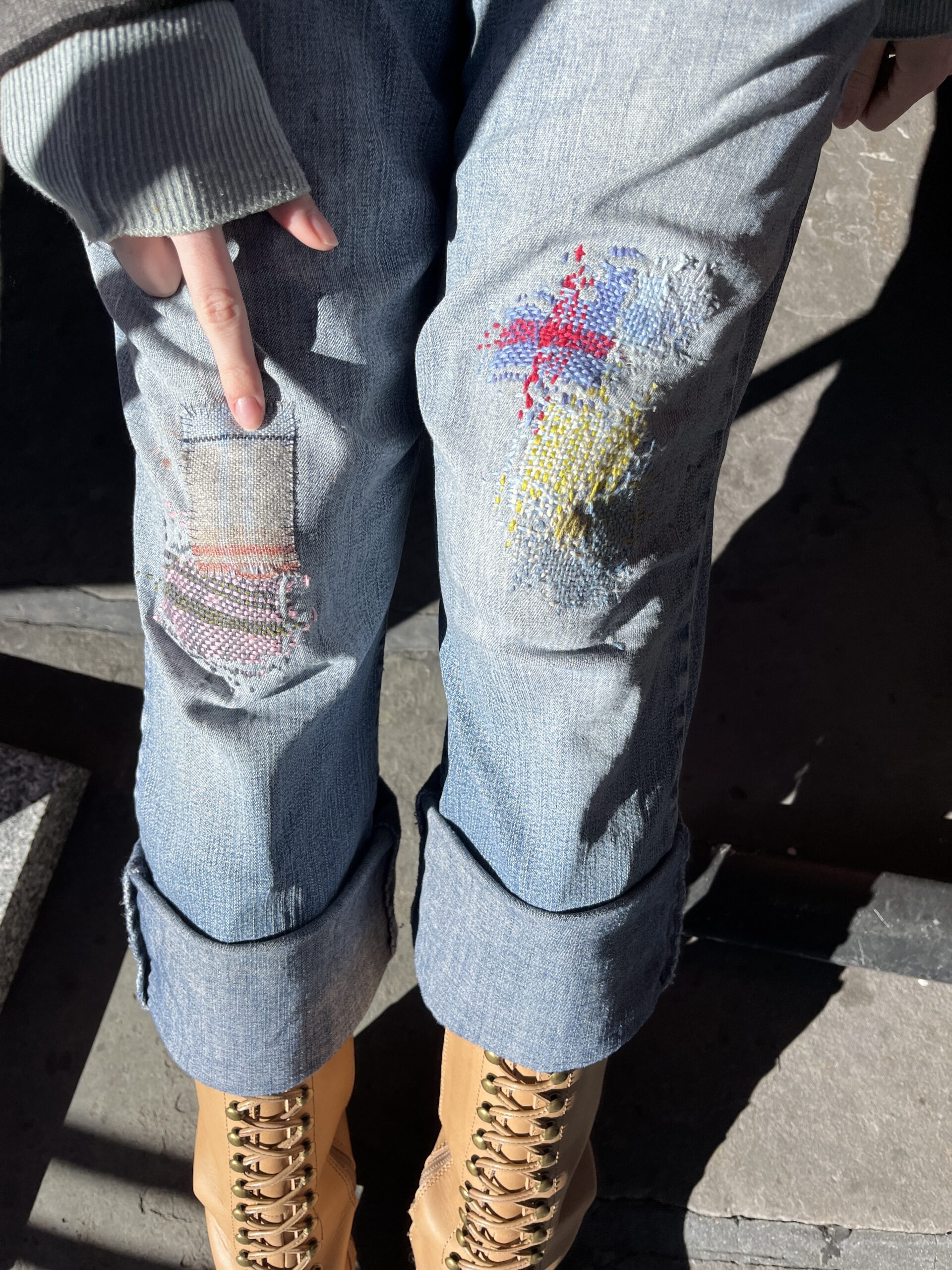 A person is wearing light blue, slightly distressed jeans with visible patches and colorful embroidery near the knees. One hand is pointing to a patch on the left knee area. The jeans are rolled at the cuffs, revealing tan boots with gold laces. The background is a stone surface with light and shadow contrasts.