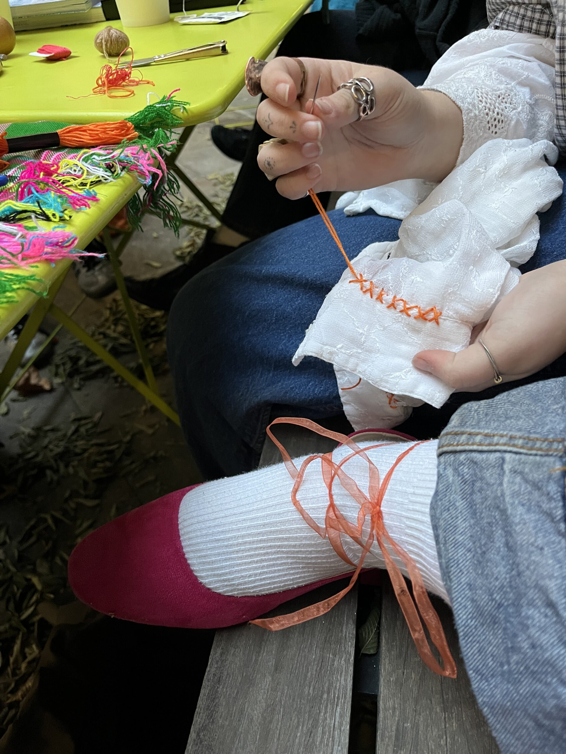 A close-up of a hand holding a needle and orange thread as it stitches into a piece of white fabric. In the foreground, a foot is visible wearing a pink shoe and white sock, tied with an orange ribbon. Surrounding the hand are colorful yarns scattered on a bright green table.
