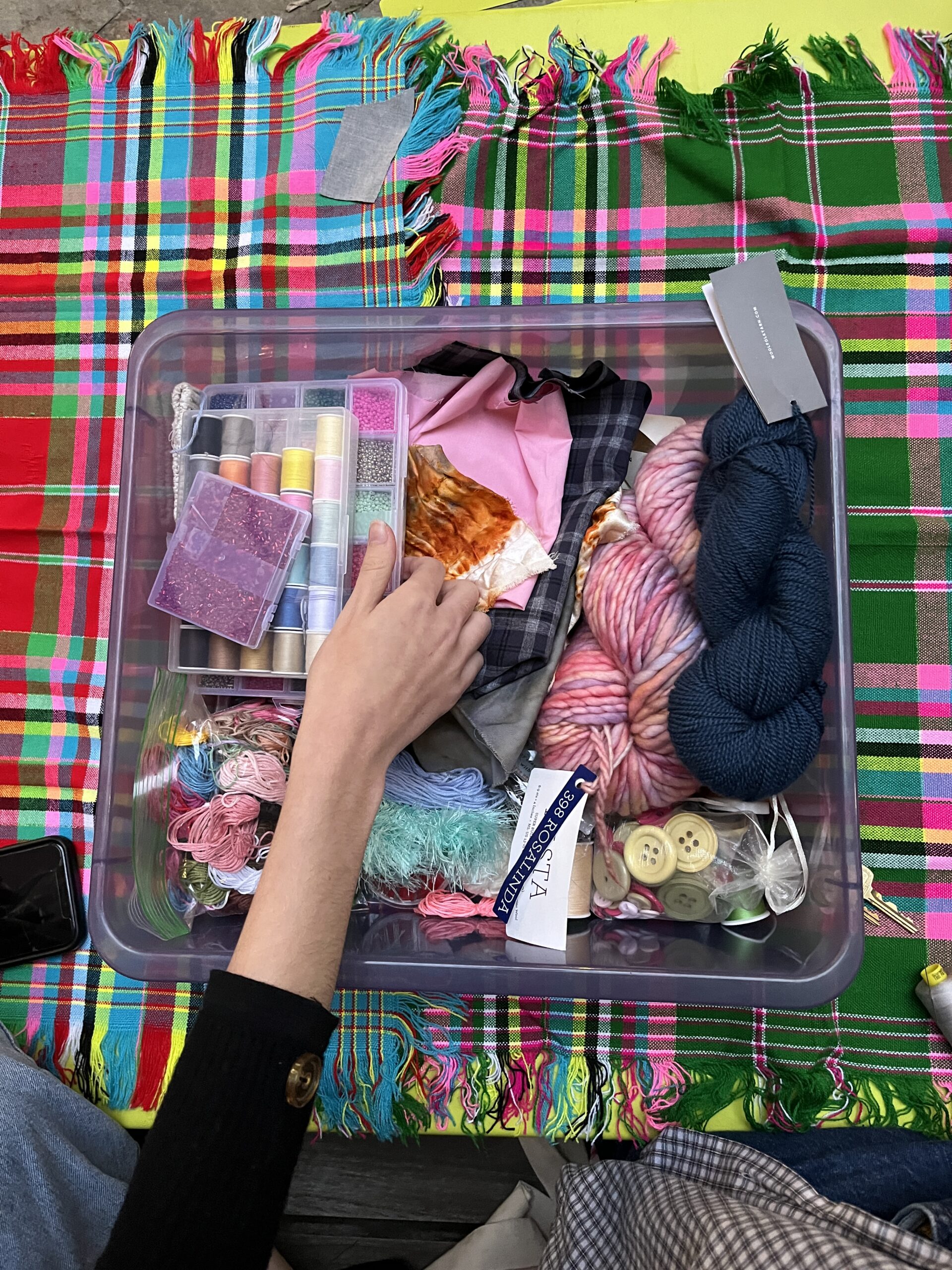 A clear plastic box is filled with various crafting supplies, including rolls of thread, beads, fabric pieces, and yarn. A hand reaches into the box to sort through the materials. Colorful woven fabrics with a checkered pattern serve as the background, showcasing vibrant hues of pink, green, and blue. The scene captures a creative workspace with a focus on textile and craft items.