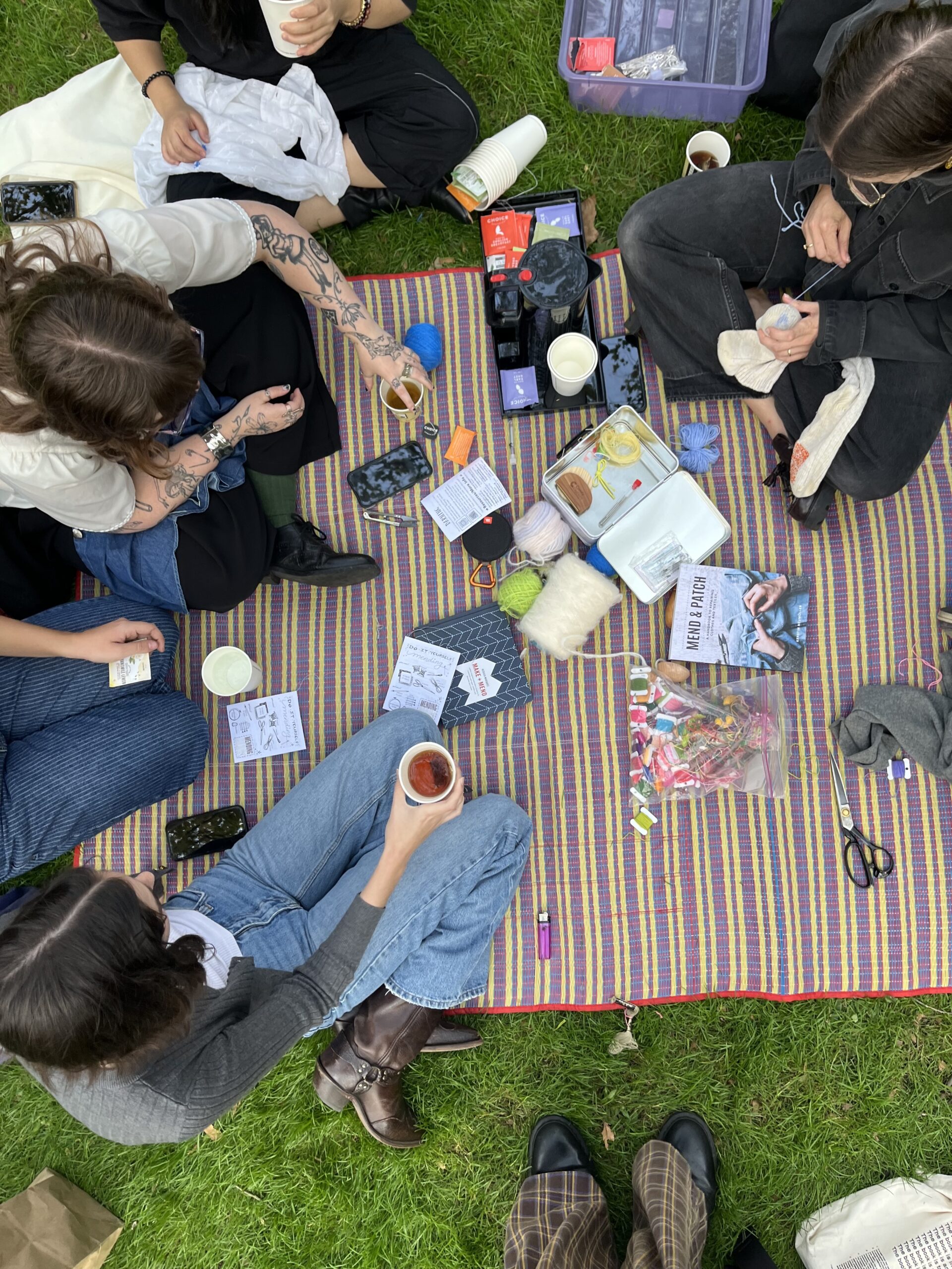 A group of people is seated on a colorful striped blanket in a park, surrounded by various craft supplies. Some individuals are working on their projects, while others hold drinks. Items visible include balls of yarn, scissors, a book, and containers with craft tools. The grass is green, and some feet in different footwear styles are visible at the bottom of the image.