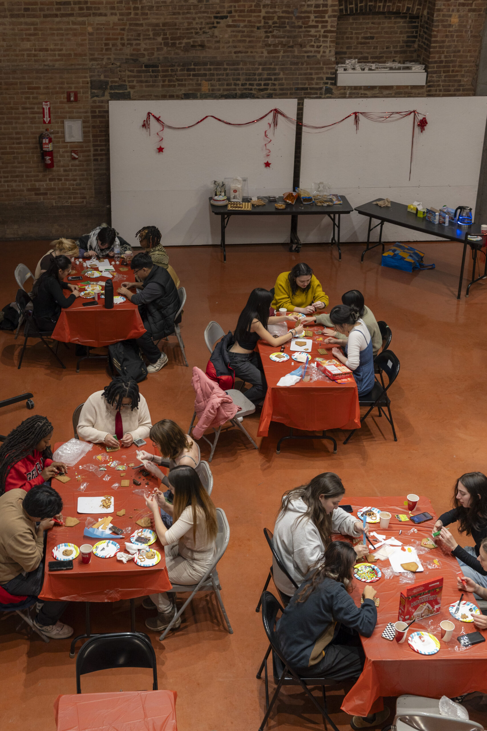 An overhead view of a large room with multiple tables covered in orange tablecloths. People are engaged in a festive activity, decorating gingerbread cookies and crafts using various colorful materials. Some participants are seated at tables while others stand nearby. Decorations like red streamers and stars are visible on the walls, and tables in the background hold snacks and supplies. The atmosphere appears social and creative.