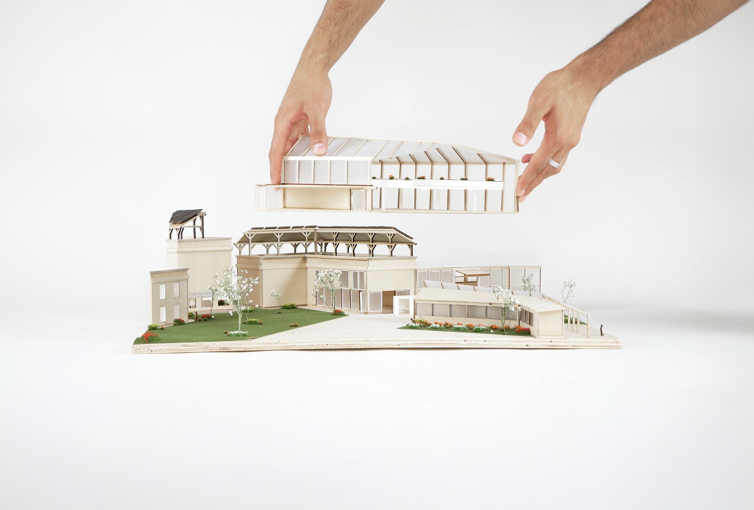A pair of hands is positioned above a detailed architectural model. The model features multiple buildings with various roof styles, greenery, and landscaping elements like grass and flowers. One hand is lifting a top component of the model, revealing the structure beneath. The background is plain white, emphasizing the model's features.