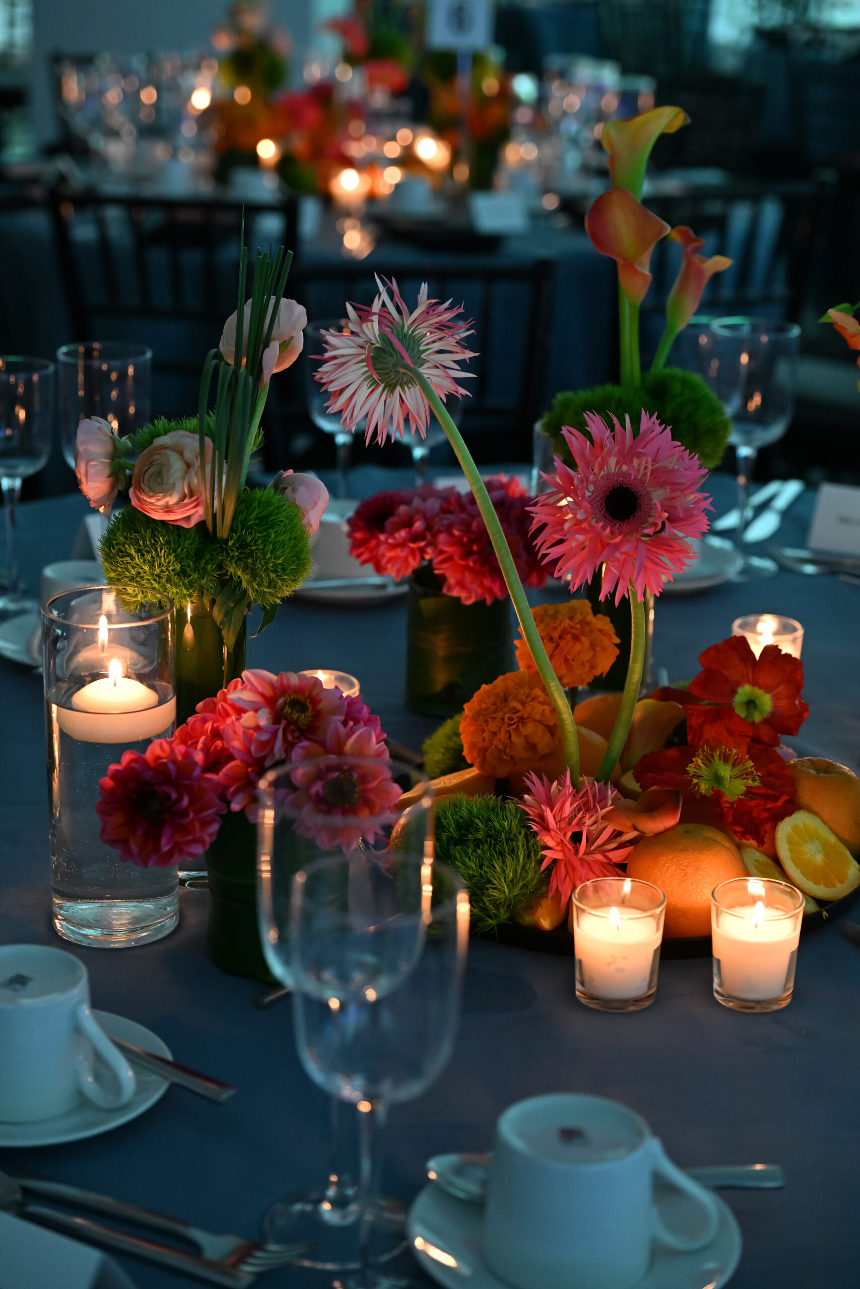 A beautifully arranged table featuring vibrant floral centerpieces in various shades of pink, orange, and green. Candles in glass holders provide soft lighting. The table is set with elegant glassware and white cups on saucers. The background blurs, revealing more floral arrangements and a sophisticated dining atmosphere.
