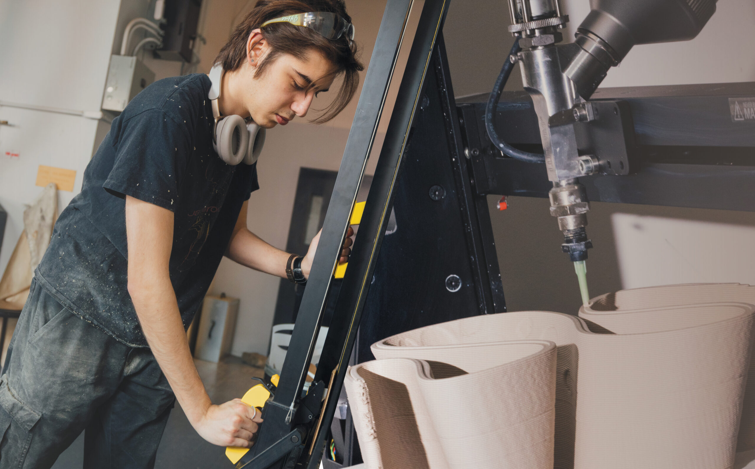 A student is adjusting a large black machine in a workshop, wearing headphones and a T-shirt dusted with particles. On the right side, a close-up of a robotic tool extruding clay into a wavy shape. The background shows a workspace filled with various materials and equipment.