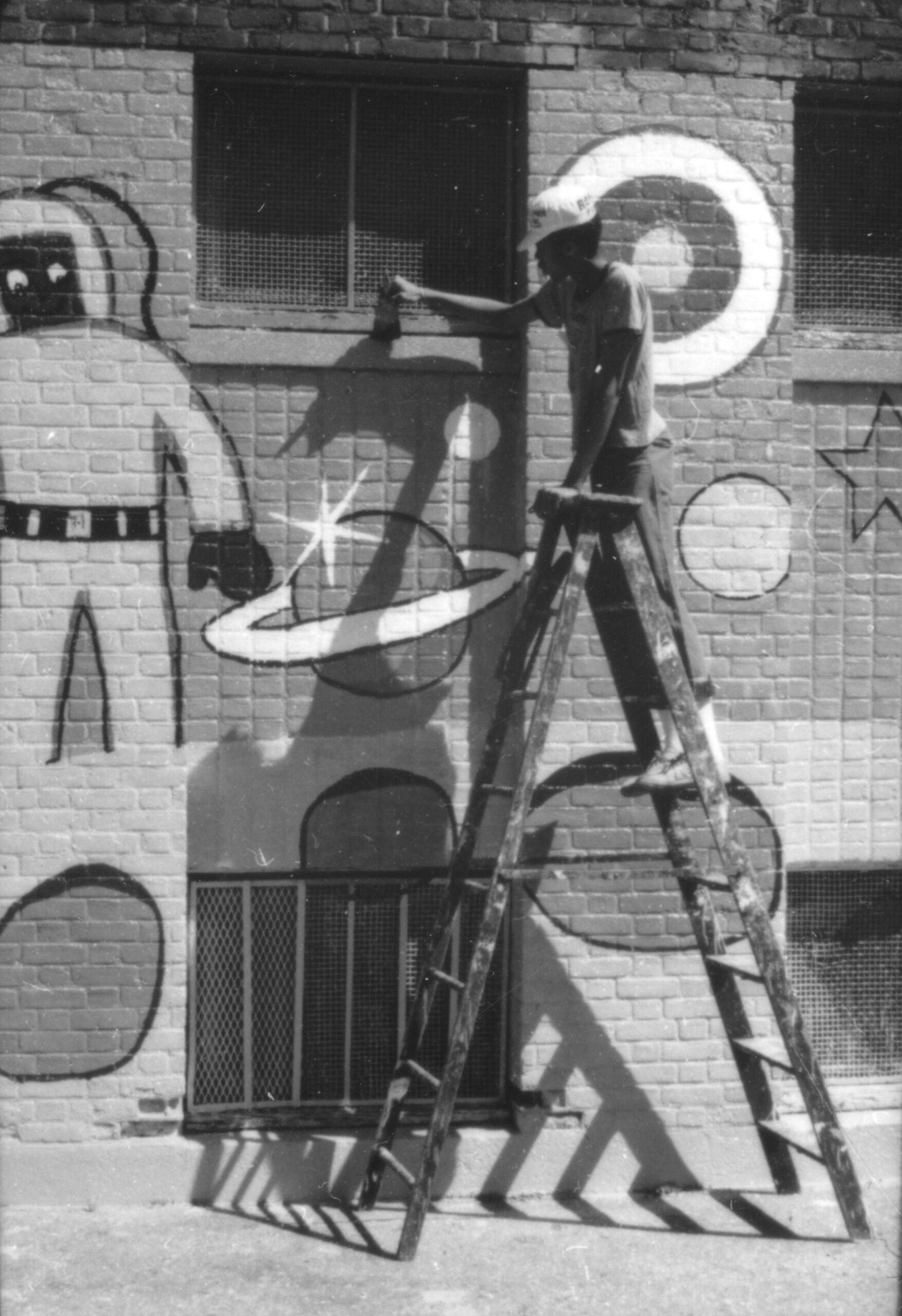 A student stands on a ladder and paints a mural, which includes an astronaut and planets.