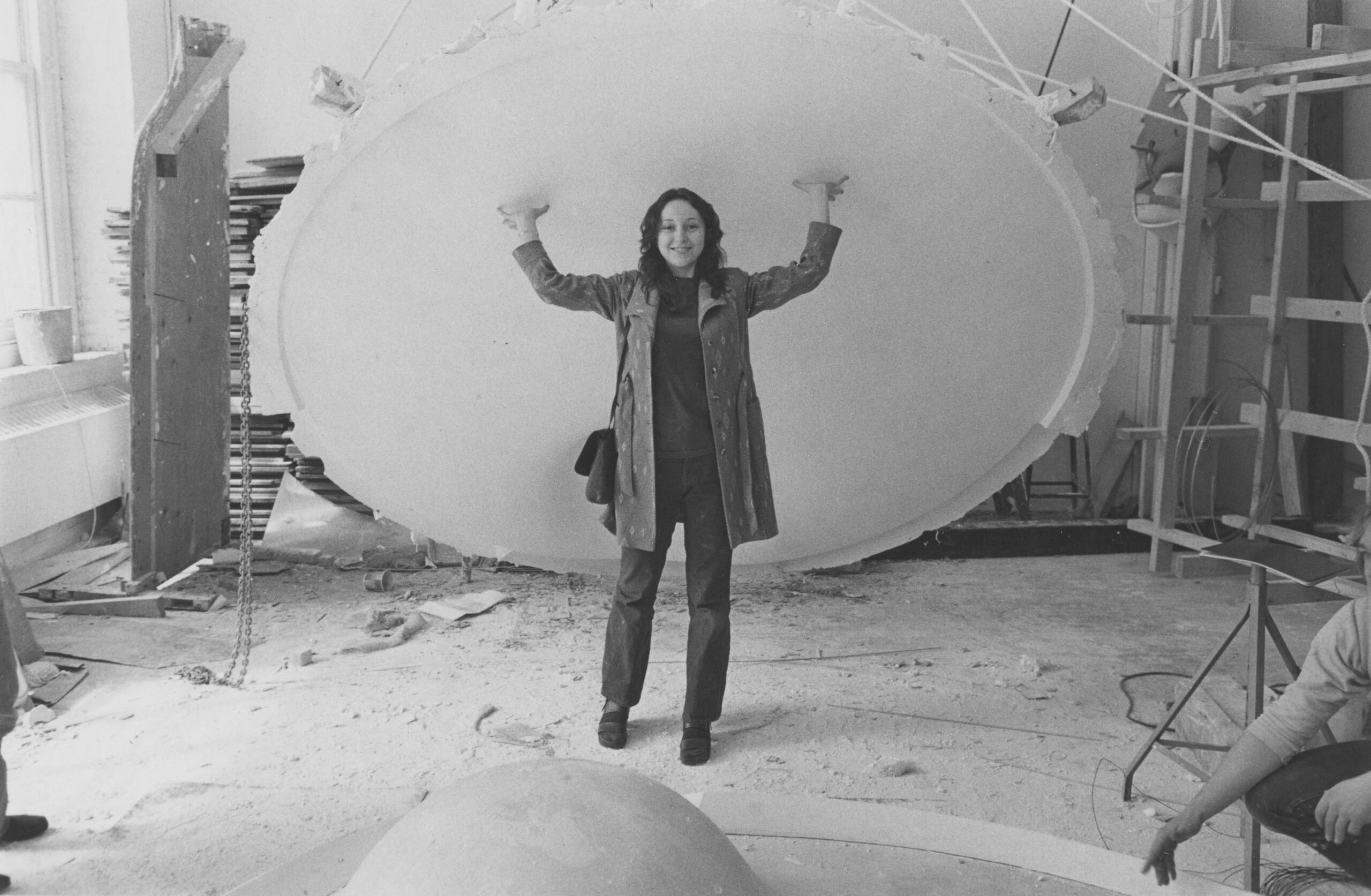 A student stands underneath a large round piece of material suspended by ropes in a studio, holding it with their fingertips and smiling, as another student crouches in the foreground.