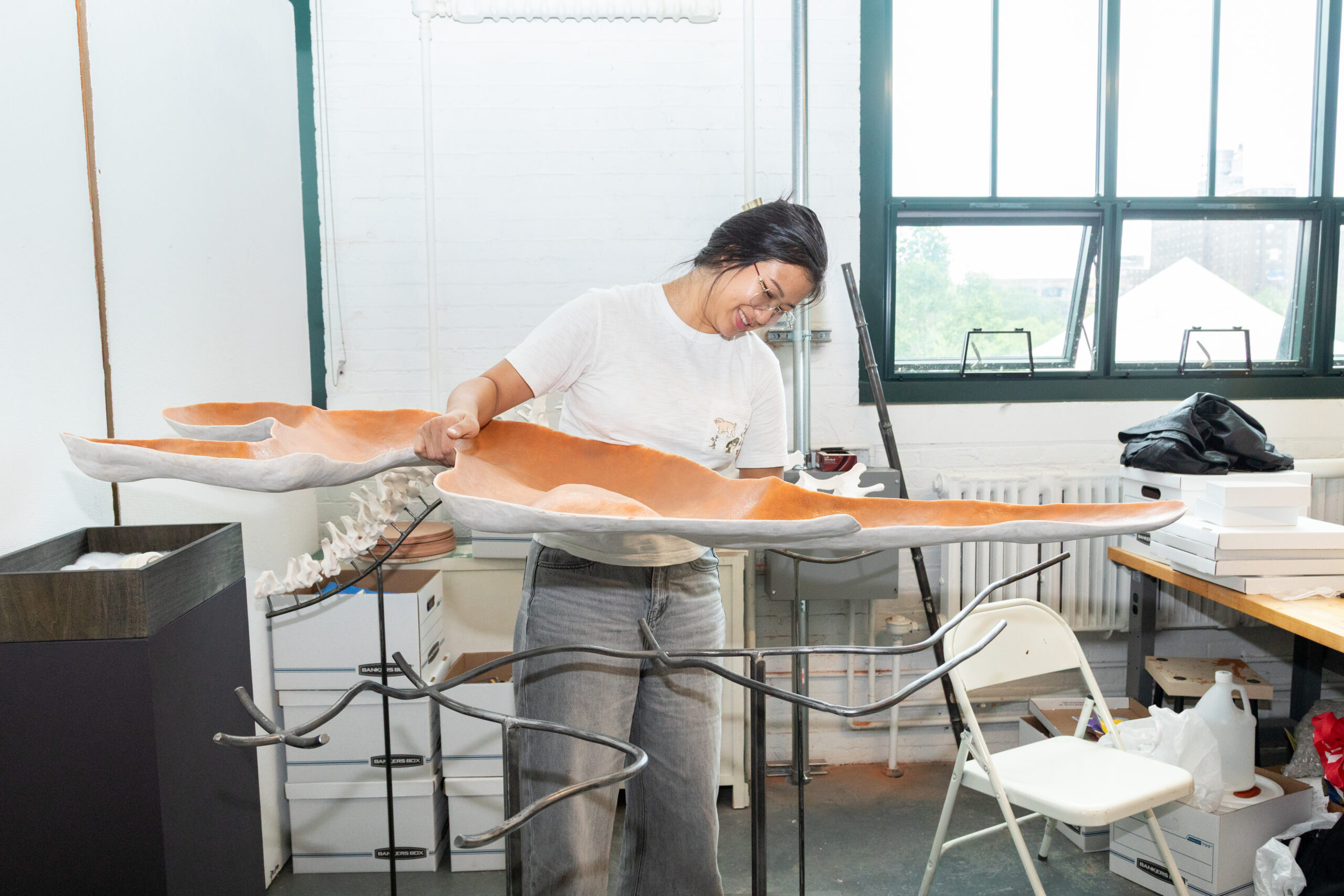 An artist, wearing glasses and a white shirt, smiles as she holds a large, sculptural piece with an orange and white color scheme, above a metal stand. The background shows a well-lit studio with various art materials, including a table, boxes, and a radiator.