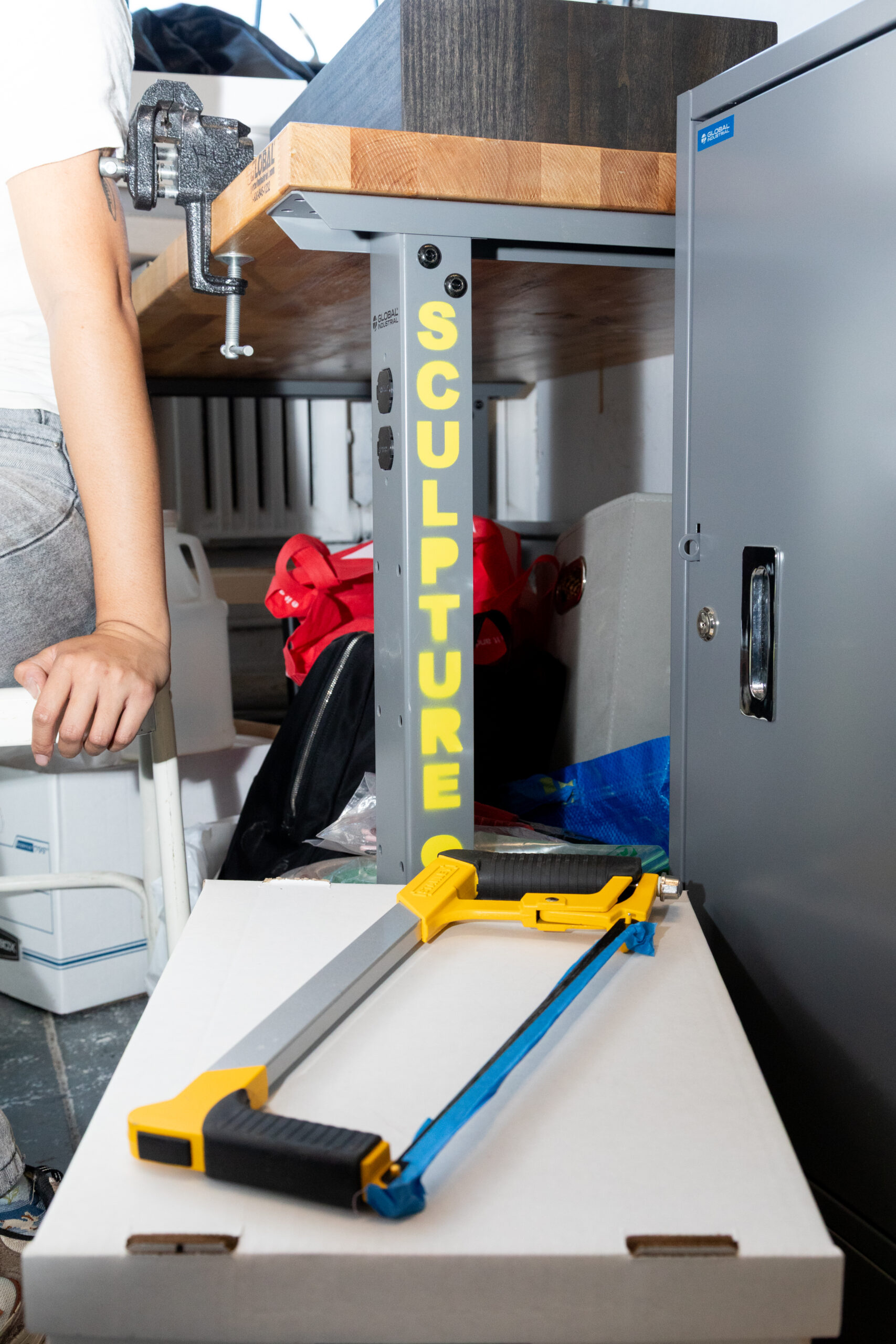 A workspace featuring a large wooden table with a metal clamp attached. The table leg is marked with yellow letters spelling "SCULPTURE." In front, there is a box, and on top of it rests a saw with a bright yellow handle. Various objects and bags are visible in the background, along with a gray cabinet. A person's hand is partially seen resting on the table.