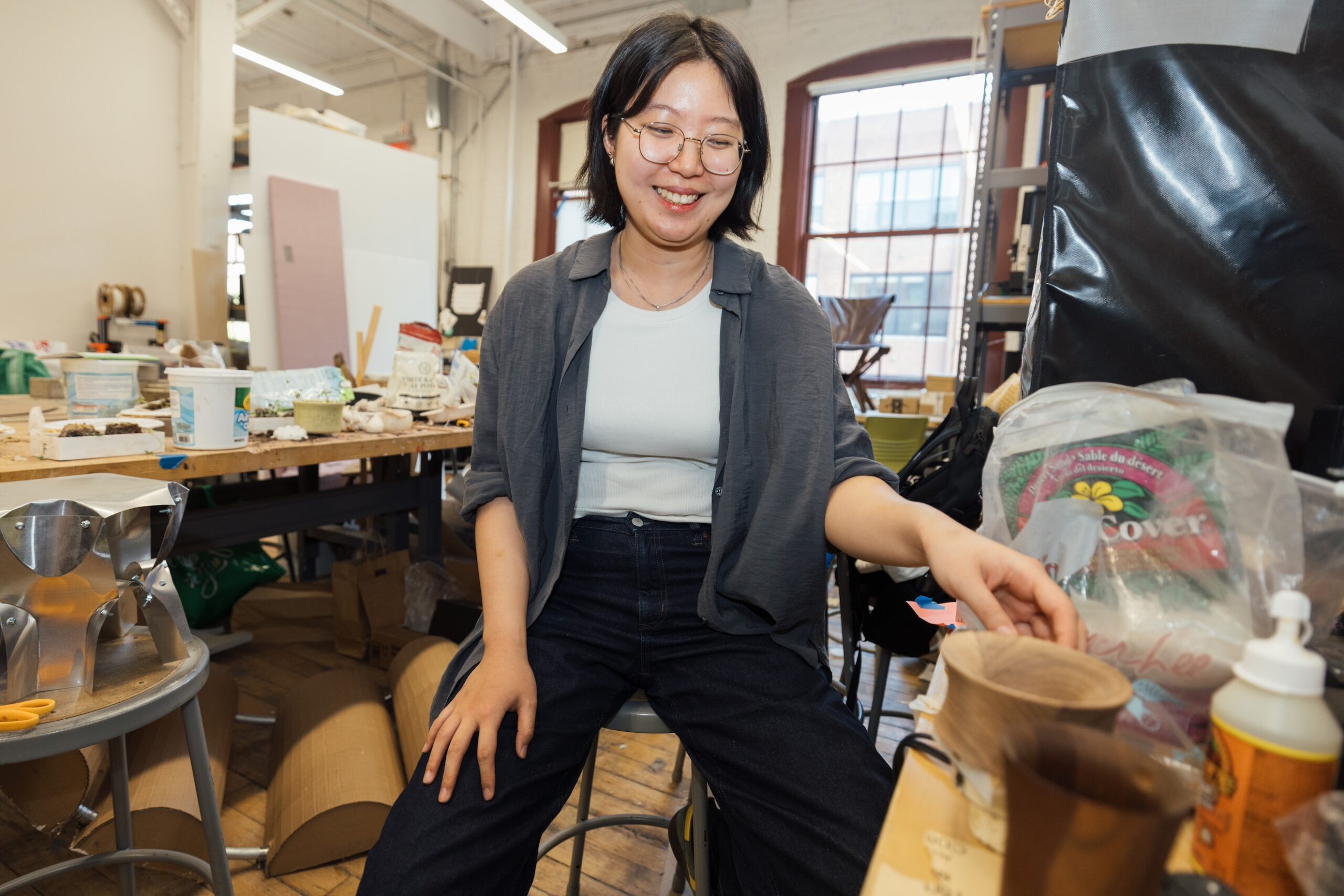 A smiling individual with short black hair and glasses sits on a stool in a workshop. Their right hand is reaching towards a small wooden bowl on a work table filled with various art supplies and materials. Large windows in the background allow natural light into the space.