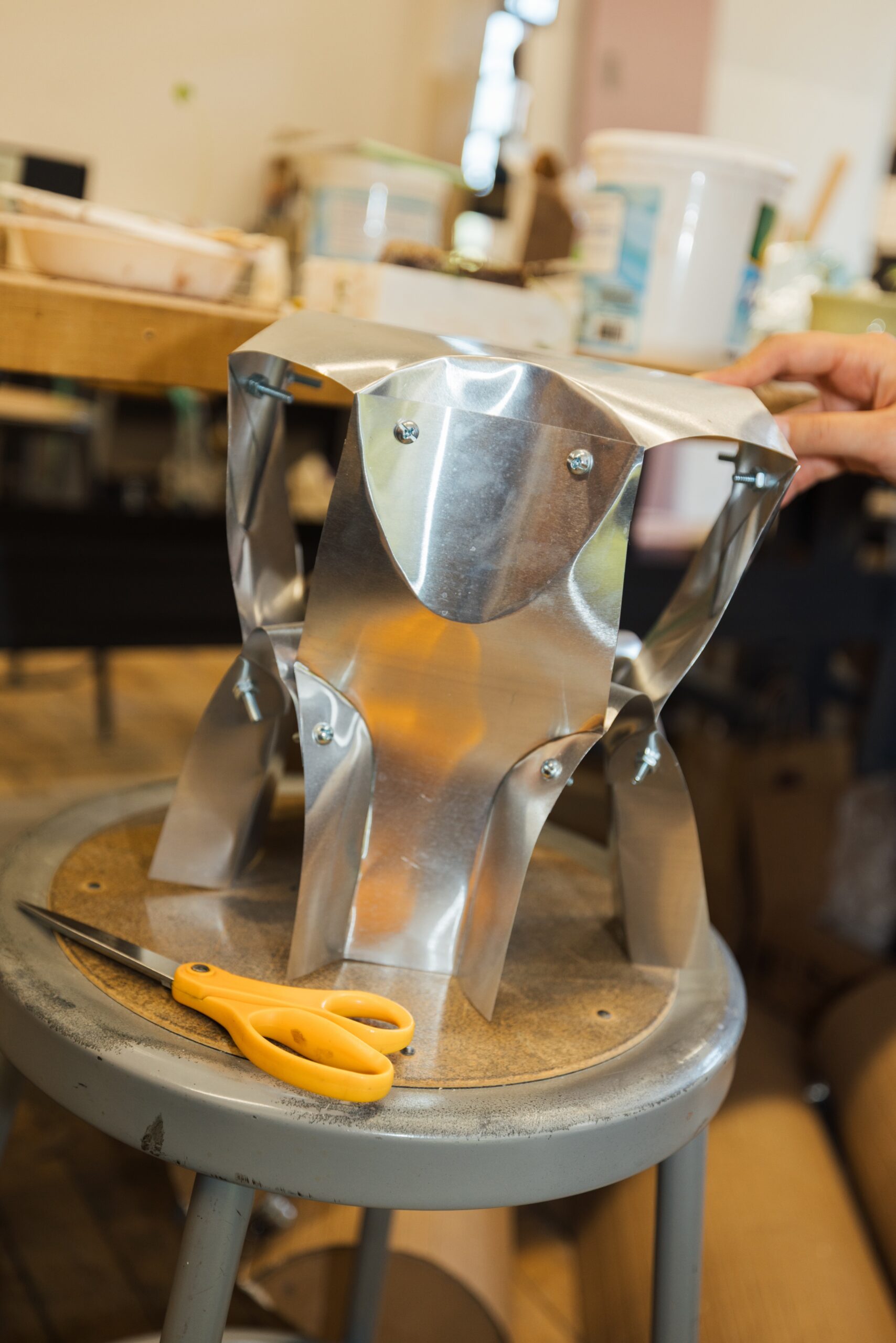 A metal structure made from shiny, curved pieces is placed on a circular studio stool. The parts are secured with screws, and a person's hand can be seen adjusting one of the pieces. A pair of orange scissors rests on the stool next to the structure. The background features various art supplies and equipment in a workshop setting.