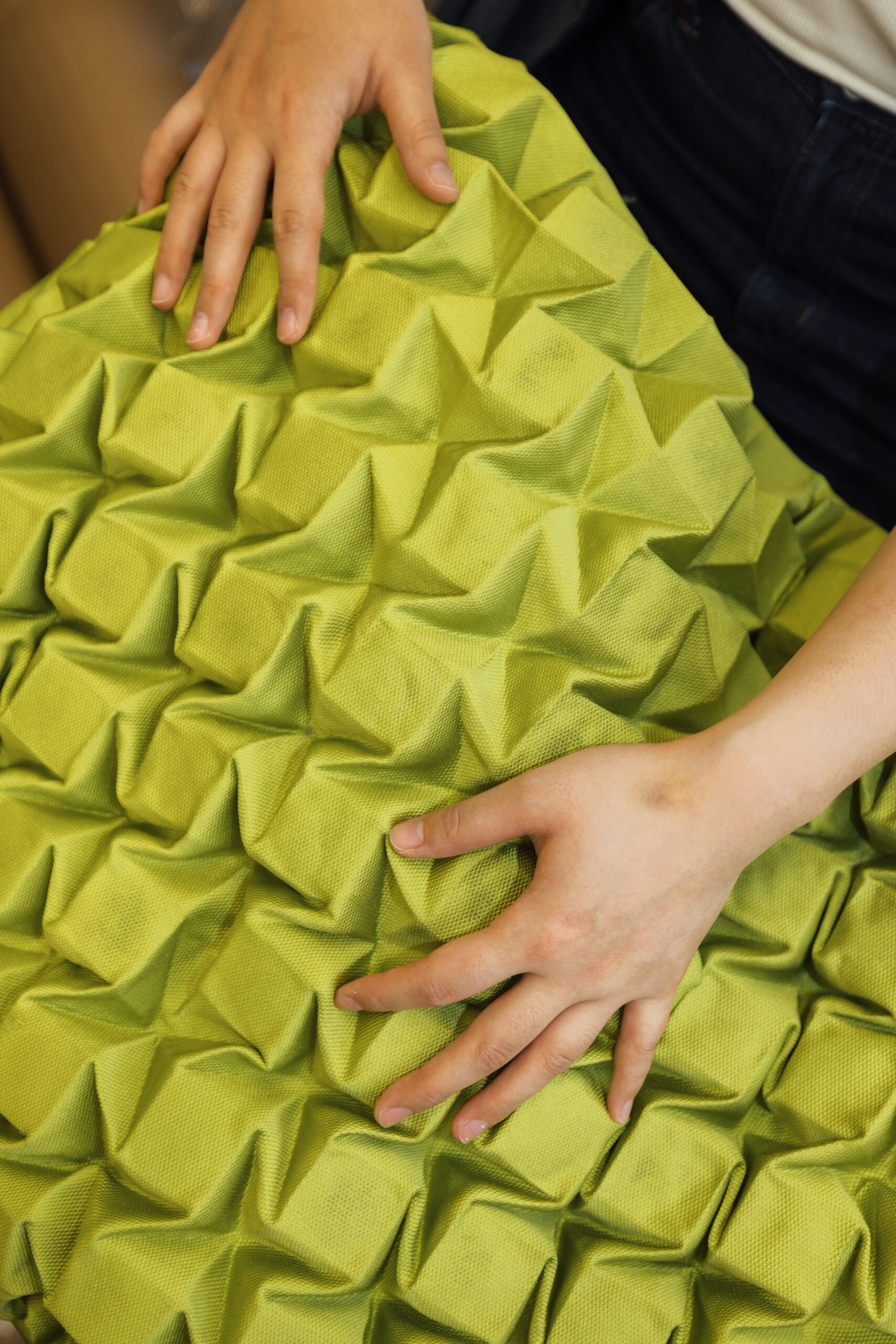 A close-up image shows two hands pressed against a textured, green cushion featuring a three-dimensional pattern of raised, triangular shapes. The fabric is soft and slightly wrinkled, emphasizing the tactile quality of the material.