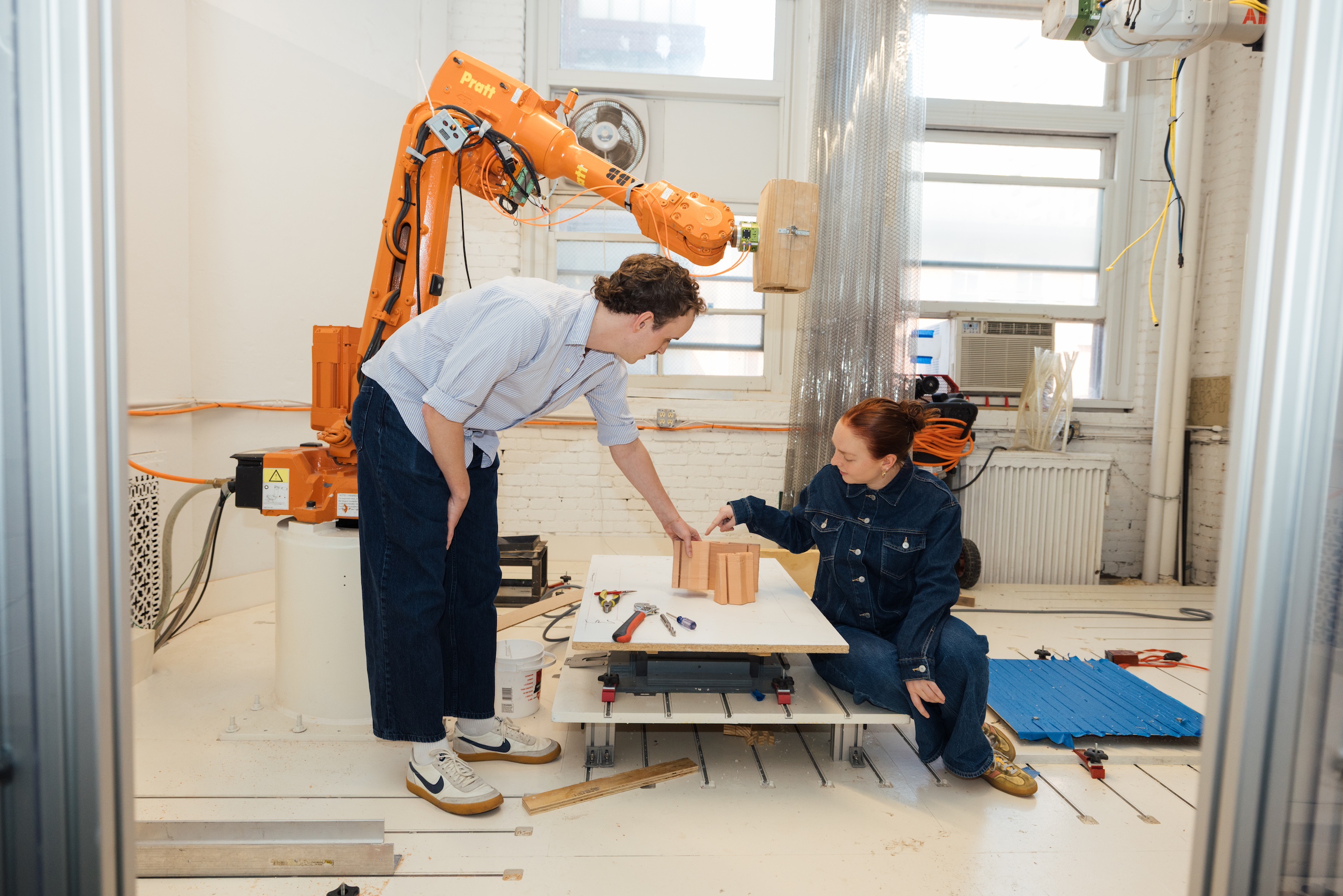 Two students working together in a workshop with a large orange robotic arm. The person on the left leans over to point at a set of clay blocks on a white table. The woman is sitting on the floor, examining the blocks. An orange robotic arm is positioned above them, and various tools are scattered on the table.
