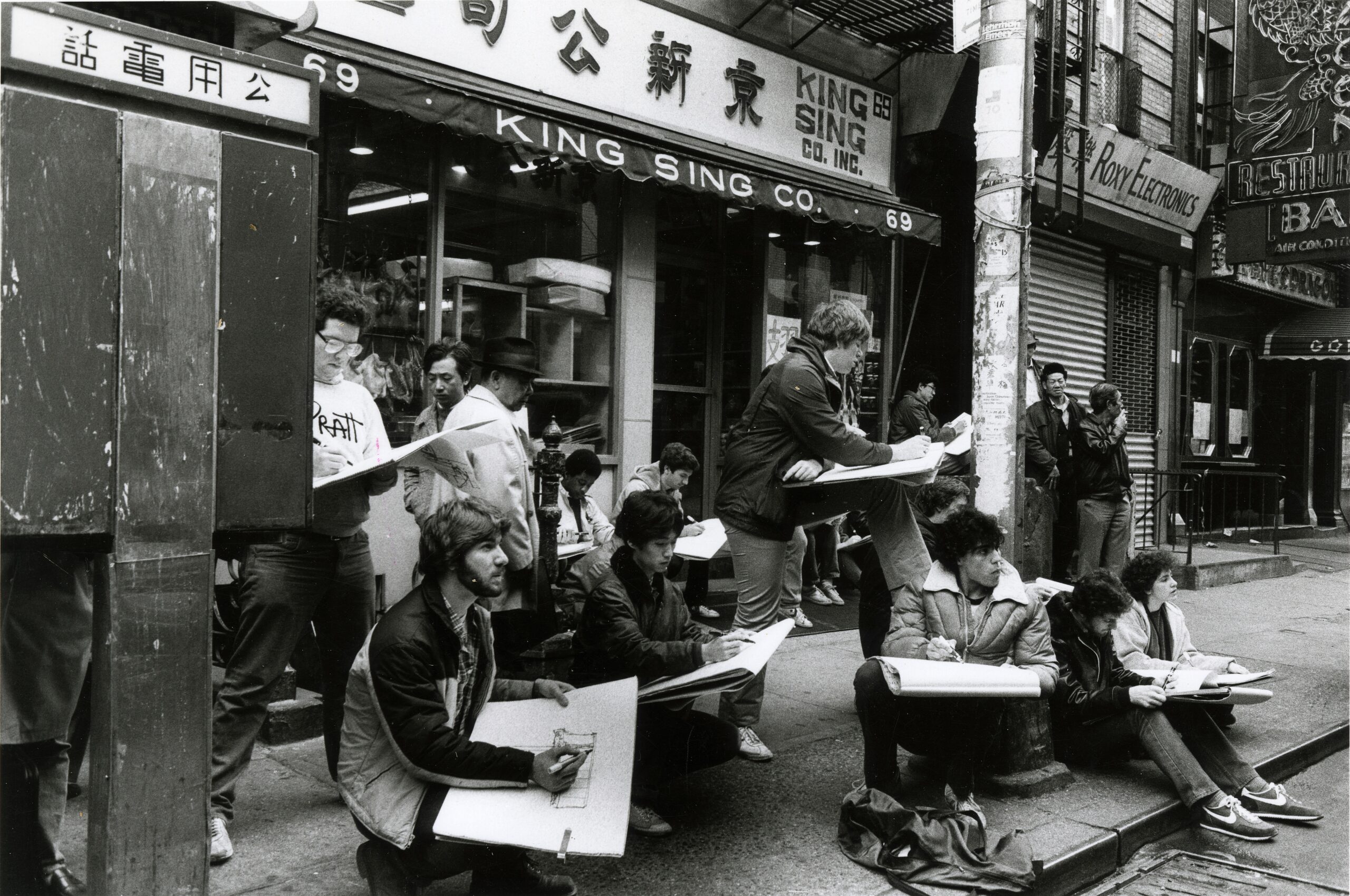 Students stand and crouch in front of storefronts in Manhattan's Chinatown while drawing something out of view across the street.