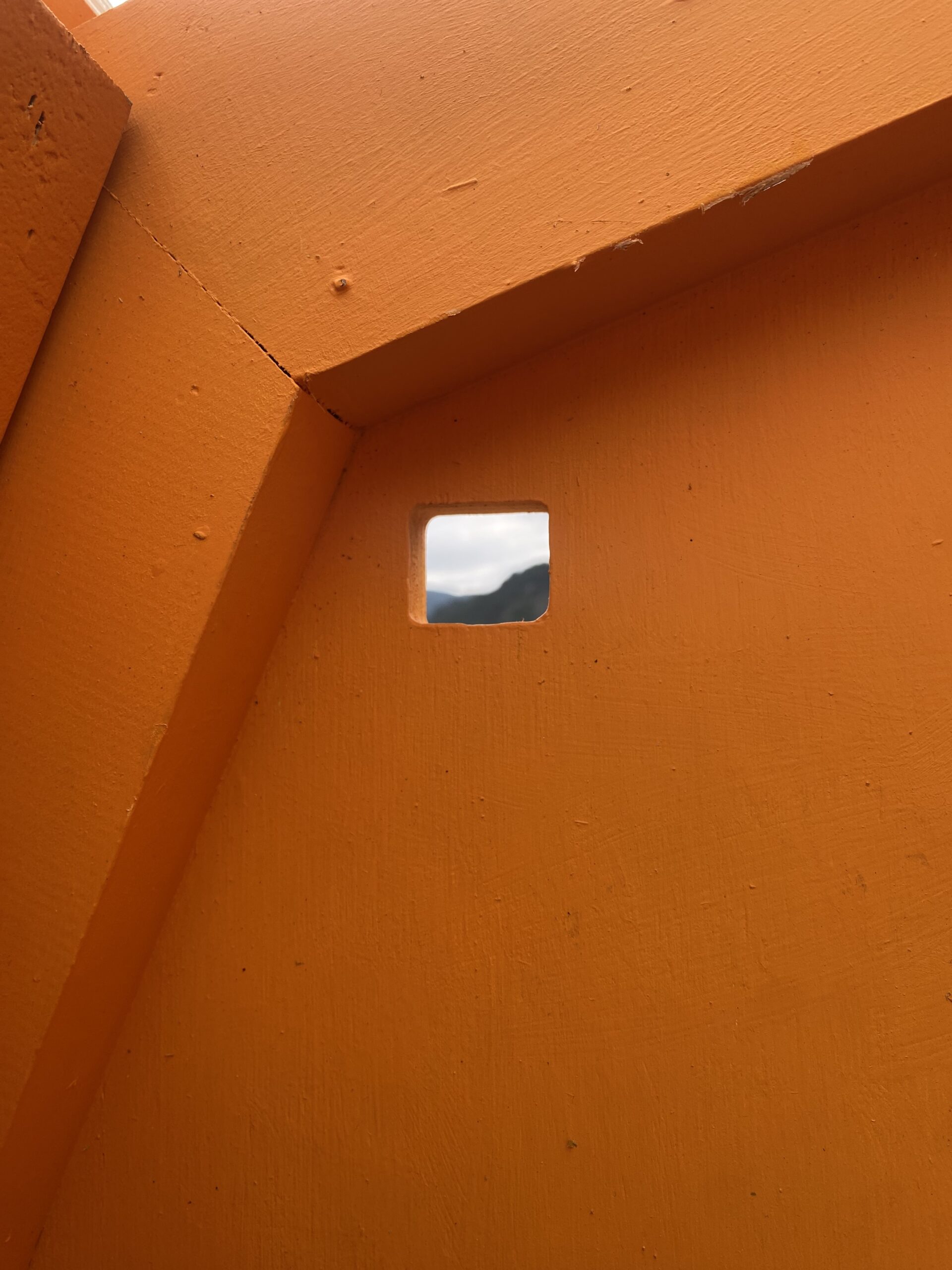 A close-up image of a vibrant orange wall featuring a small square opening. Through the opening, a view of cloudy skies and distant mountains is partially visible. The textures of the painted wall are evident, showcasing smooth and rough surfaces.