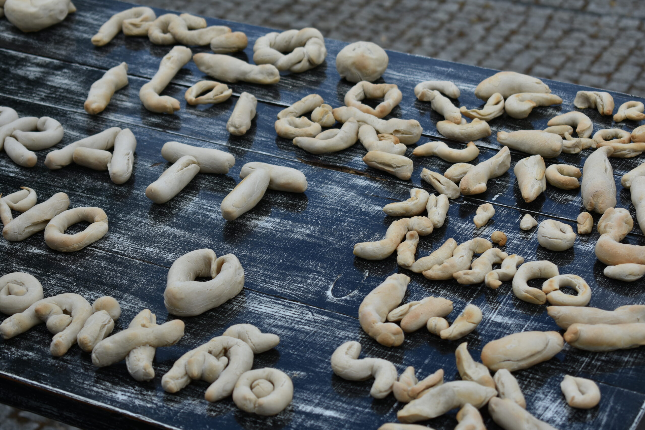 An assortment of sculpted dough shapes placed on a weathered dark wood table. The shapes resemble various letters and abstract forms, with a light beige color. The surface of the table shows a rustic finish, contrasting with the smooth, soft appearance of the dough pieces.