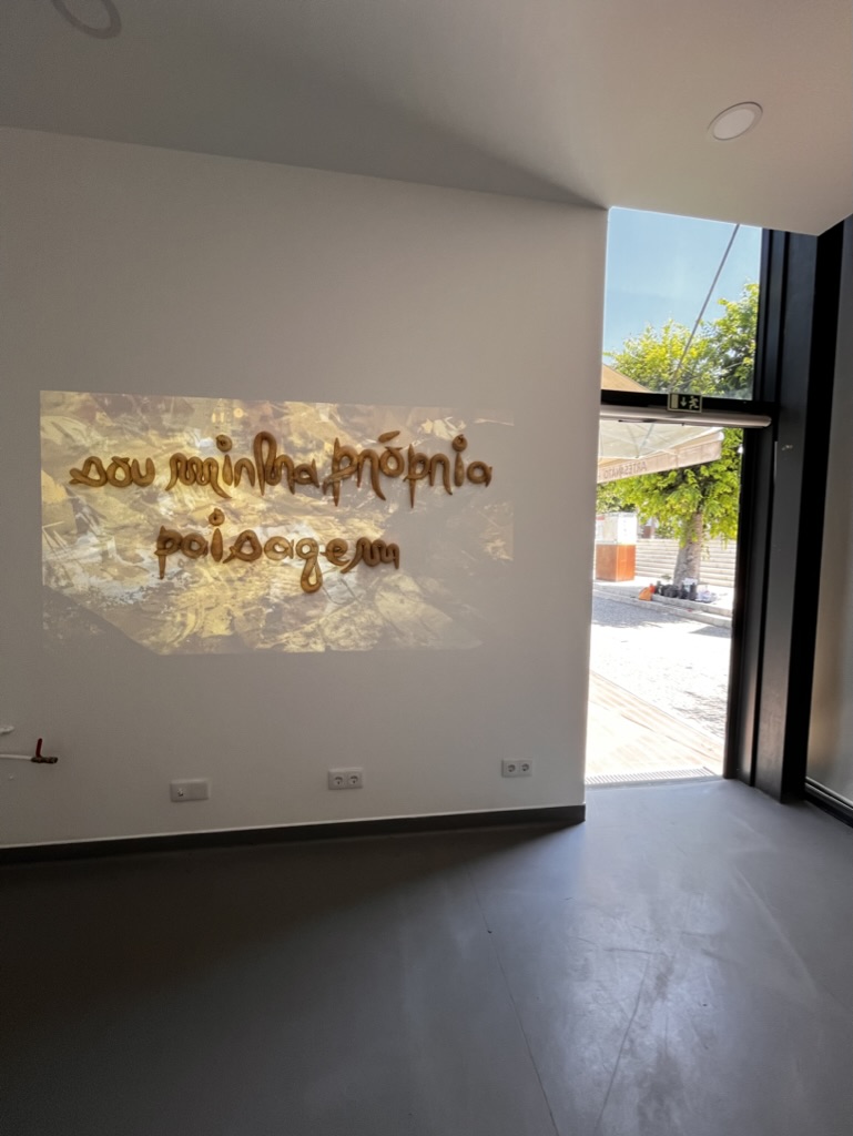 A projection on a white wall displays the phrase "sou minha própria paisagem" in a stylized, golden font made of bread. The background consists of earthy tones and textures. To the right is a glass door revealing an outdoor area with trees.