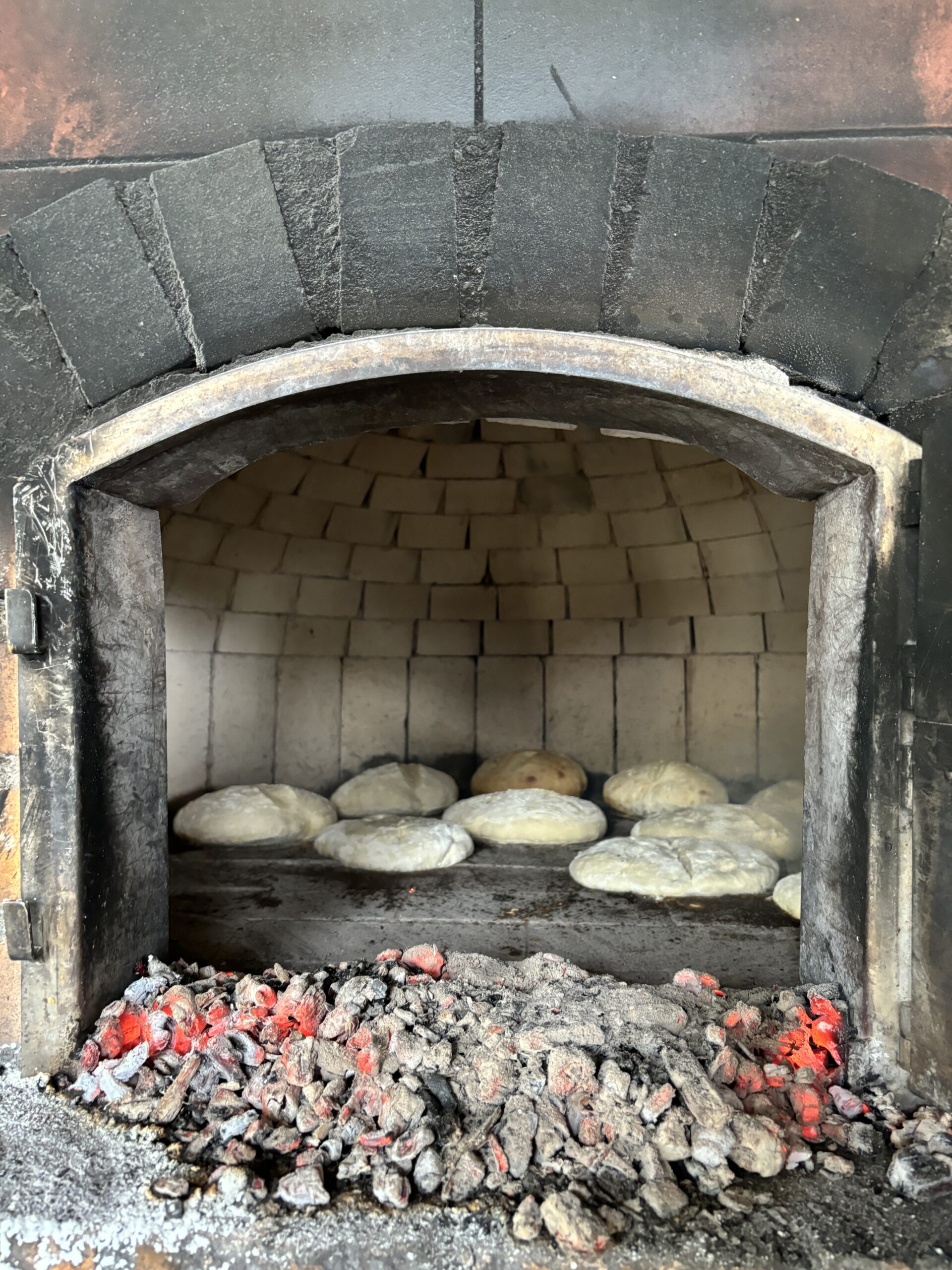 The inside of a traditional brick oven with a curved entrance. Inside, several dough rounds are resting on a stone floor, while a pile of smoldering embers and ash is positioned at the bottom, indicating that the oven is heated. The walls of the oven are made of beige bricks, creating a warm, rustic atmosphere.