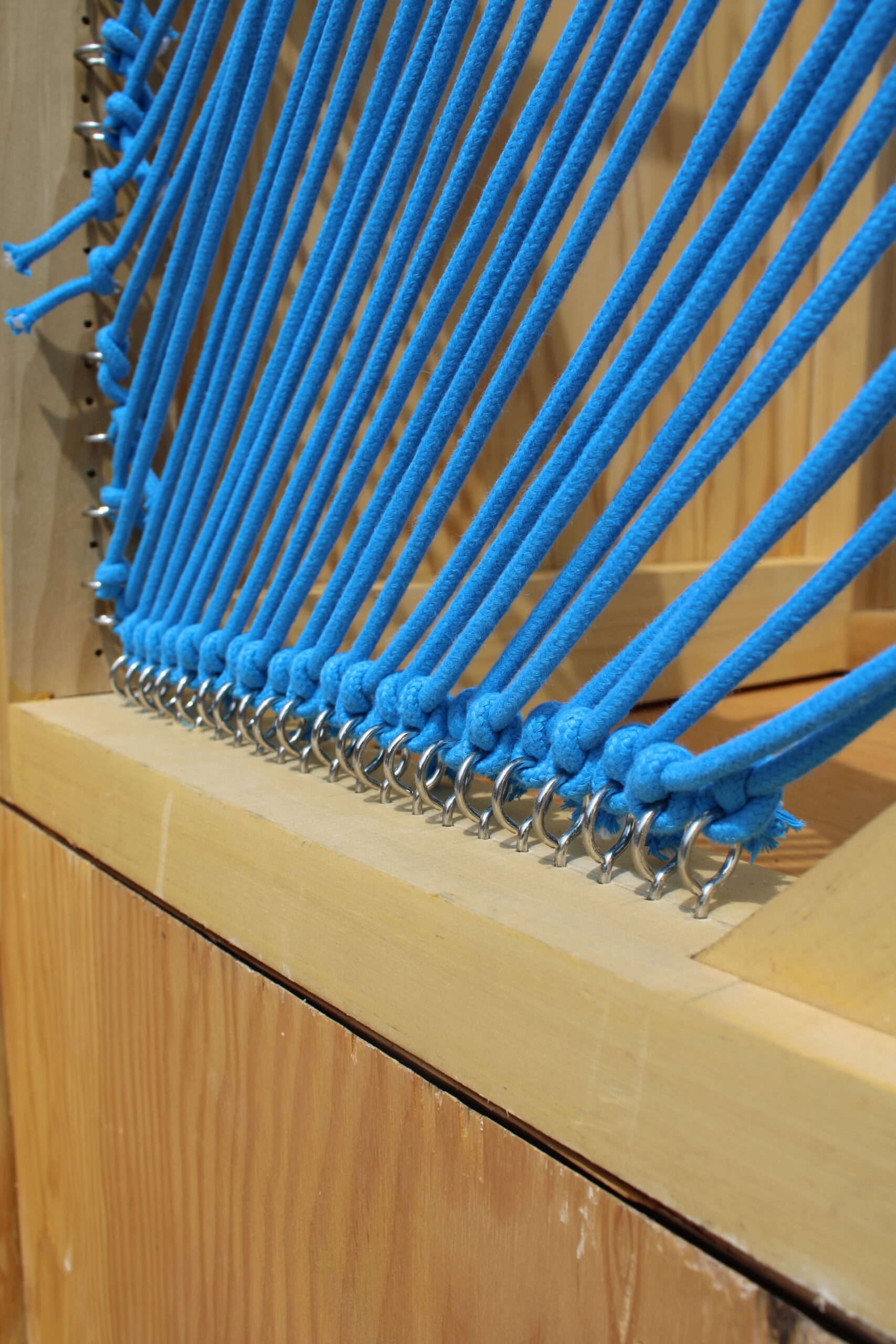 Close-up of blue ropes or cords looped through metallic rings and anchored to a wooden frame. The ropes are tied in knots at the bottom, showcasing a structured, linear pattern. The background consists of a light wood surface, emphasizing the contrast between the blue cords and the natural wood tones.