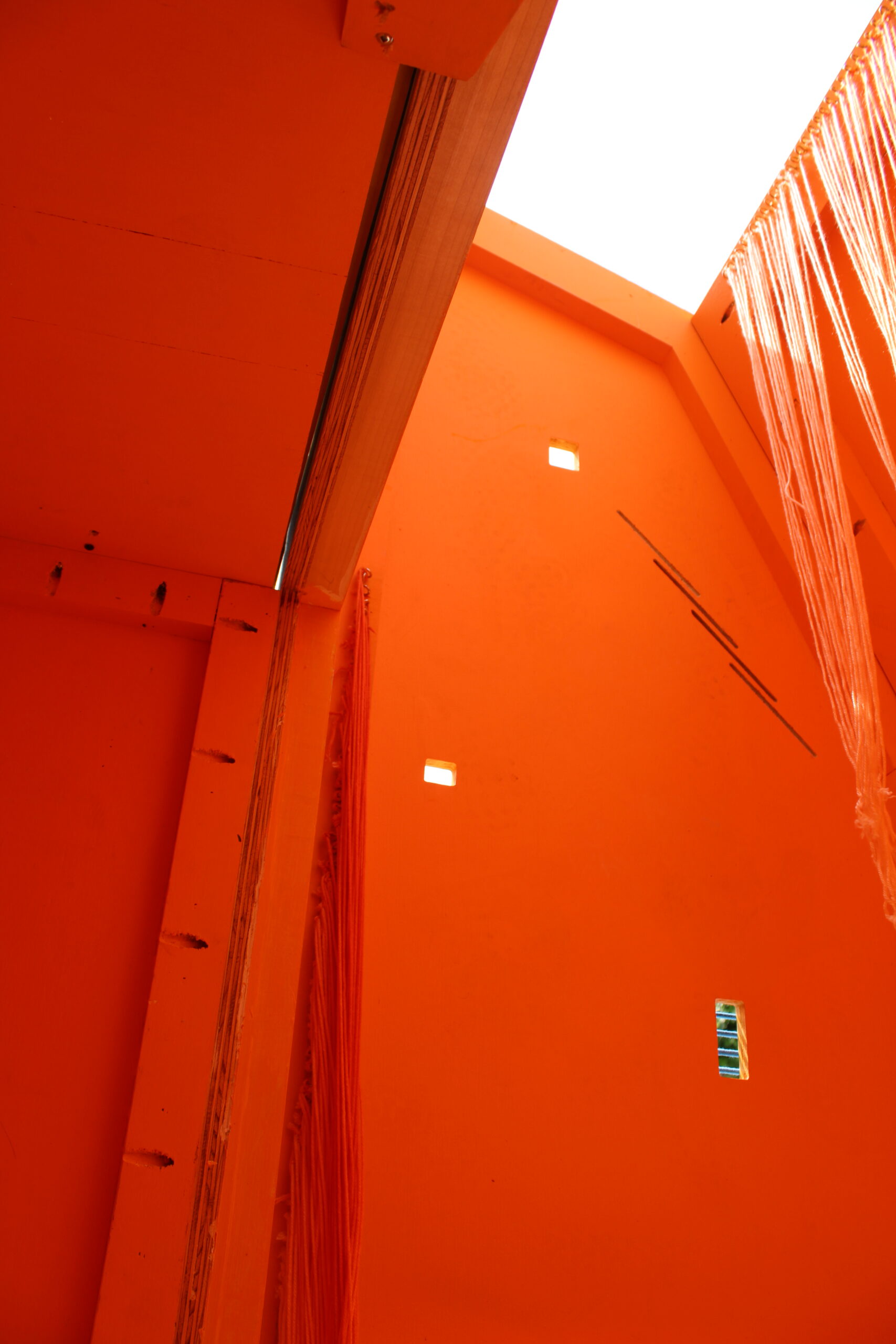 An interior space predominantly painted in bright orange. The view is from a low angle, looking up toward a skylight at the top, which allows natural light to seep in. There are two small square openings in the orange walls and strands of material hanging down, adding texture and dimension to the space.