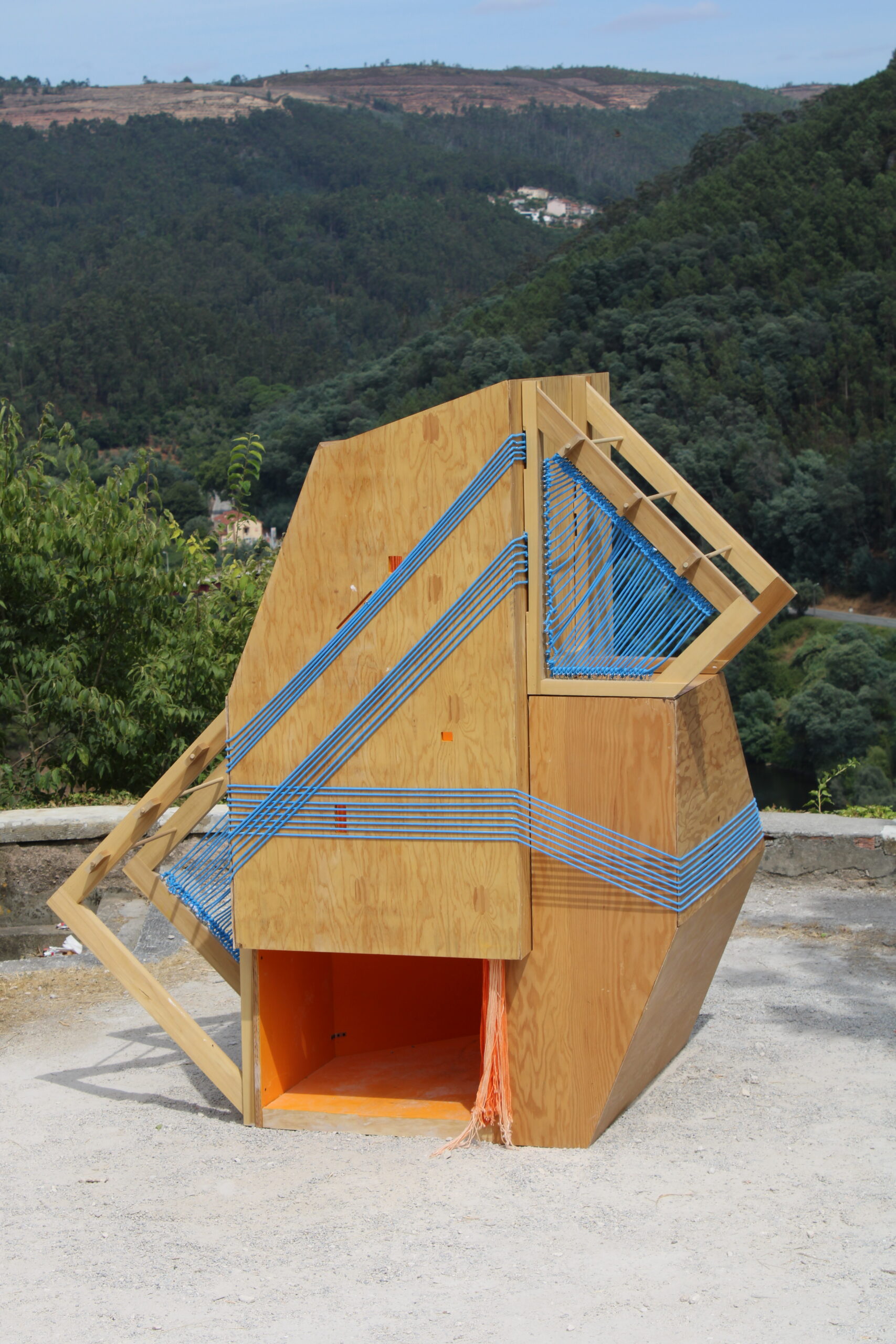 A uniquely shaped wooden structure sits on a rocky surface surrounded by green hills. The structure features a geometric design with angular sides and horizontal blue ropes woven across its surface. A small entrance is visible at the bottom, revealing an orange interior. The surrounding landscape includes trees and distant hills under a clear sky.