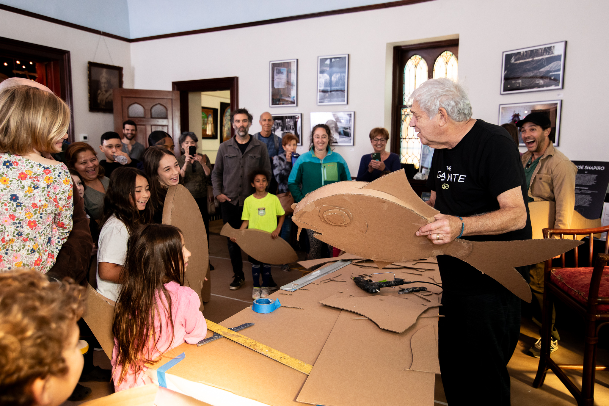 Artist James Grashow holds up a sculpted cardboard fish while interacting with a group of children and their parents, some of whom hold their own cardboard objects.