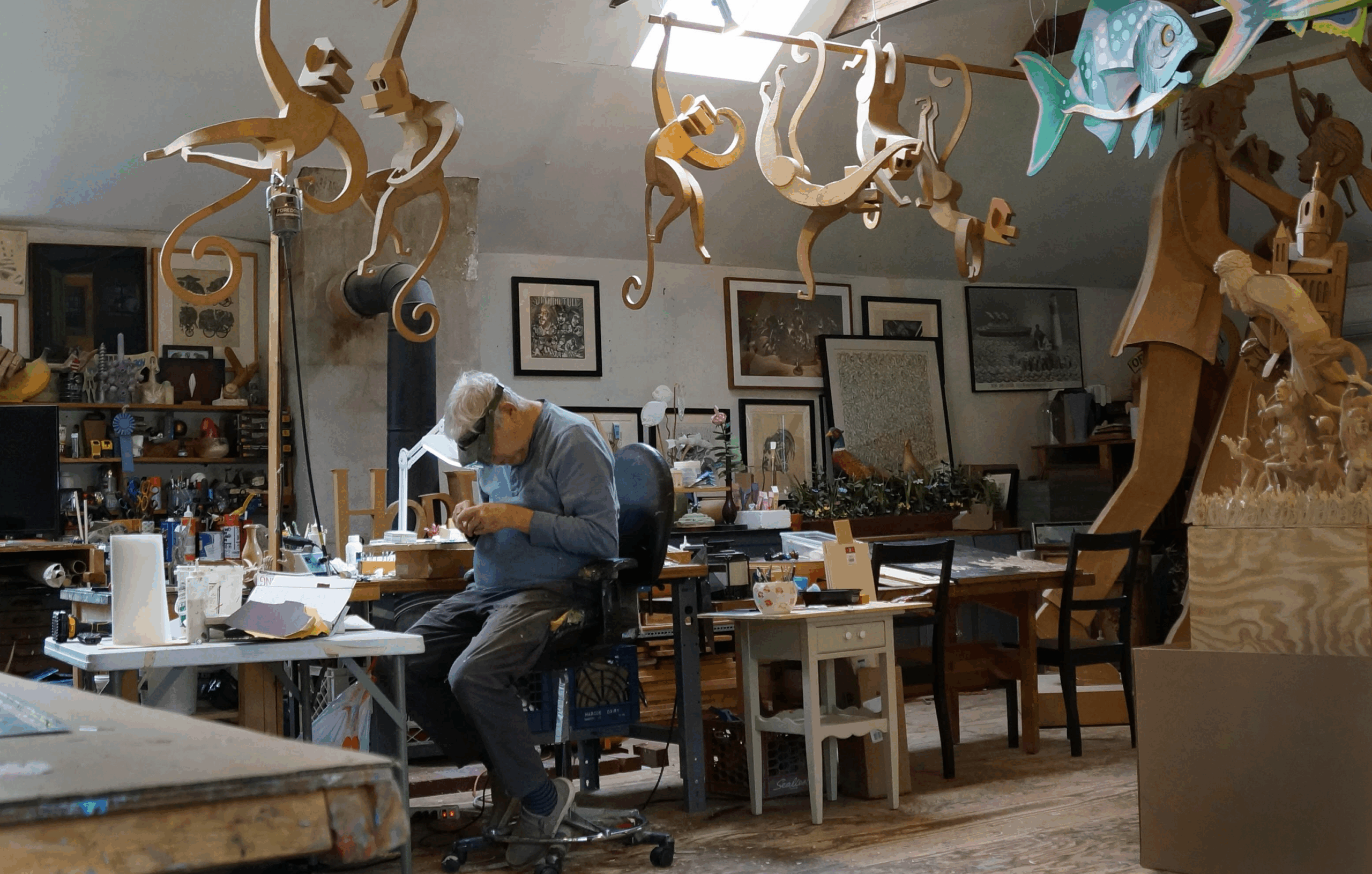 Artist James Grashow sits in a chair in his studio with his head bowed, surrounded by large cardboard sculptures of monkeys, fish, and a couple dancing.