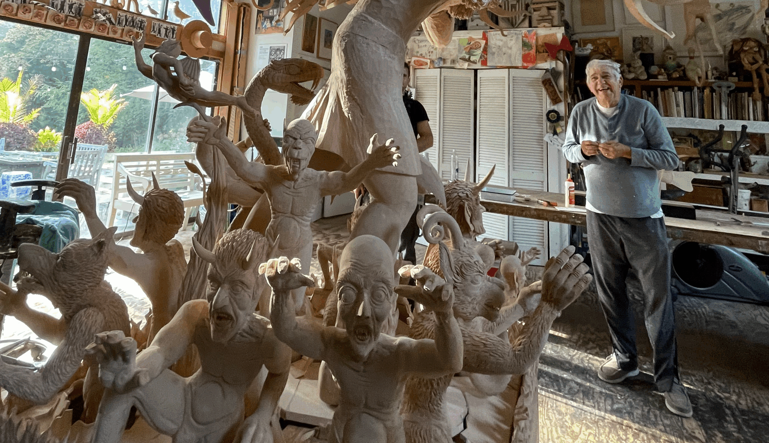 Artist James Grashow smiles in his studio behind a sculpture featuring numerous fantastical creatures.