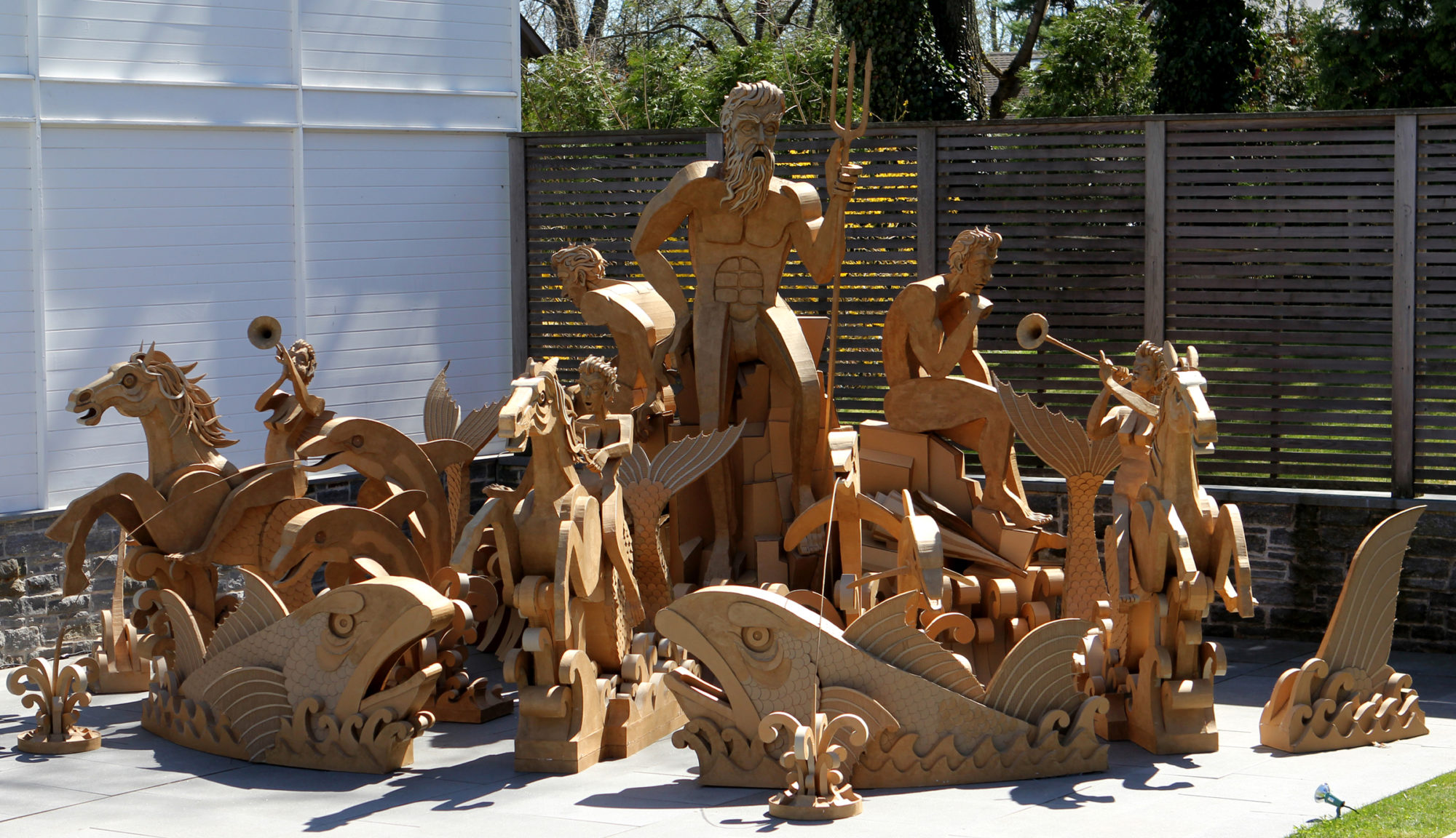 A cardboard fountain sculpture installed outdoors, featuring human figures, horses, and large fish positioned around a sea deity.