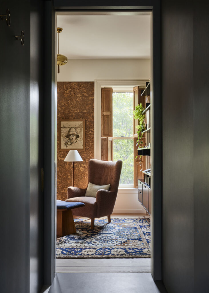 Seen through a dark gray doorway, an office with cork-lined walls. Decorating the space are a brown chair complemented with a reading lamp and footstool with a blue velvet foot rest. On the wall is a bookshelf filled with books and a vibrant green plant with dangling tendrils. A window looks over vibrant greenery.