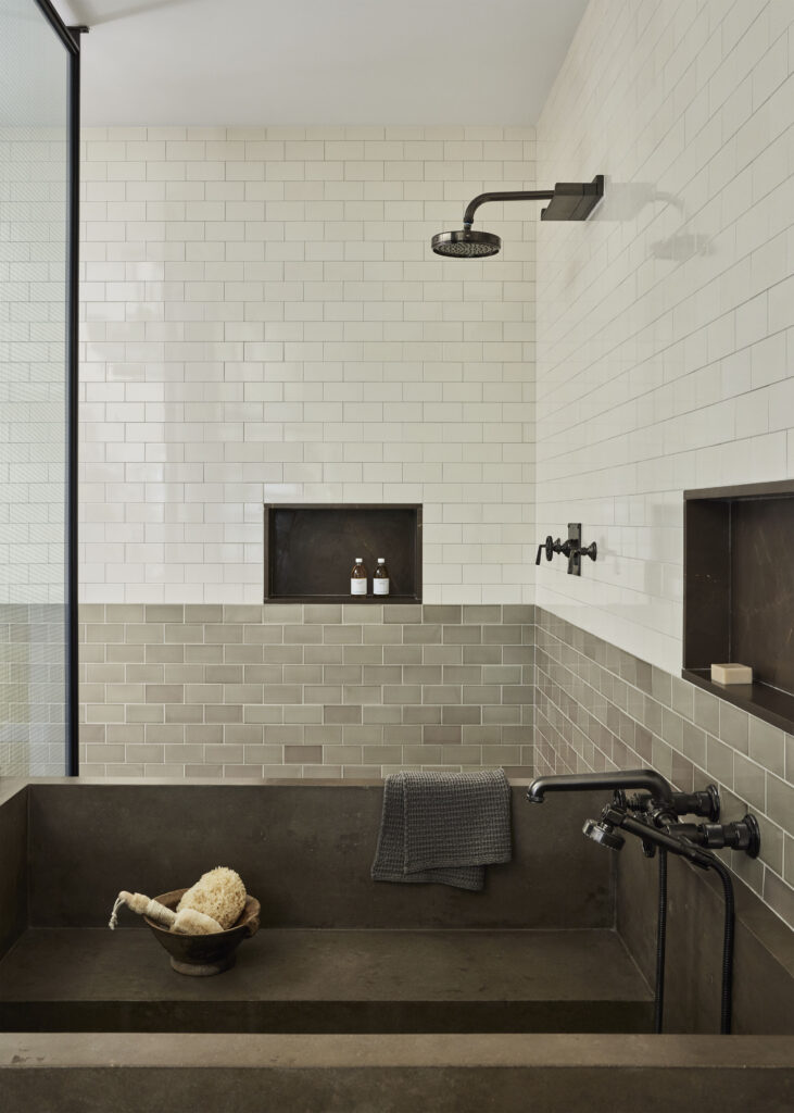 A sleek tiled shower room. The top of the shower room is lined with off-white tile. The bottom of the shower is lined with gray tile that has subtle variations in color from warm brown to sage green. The shower's fixtures, including the shower head, the faucet, and the basin are dark gray. There are two dark gray alcoves holding soap bottles and a soap bar. In the basin, there is a shelf where a wooden bowl holding nautral loofahs rest. A dark gray waffle towel hangs over the side of the basin.