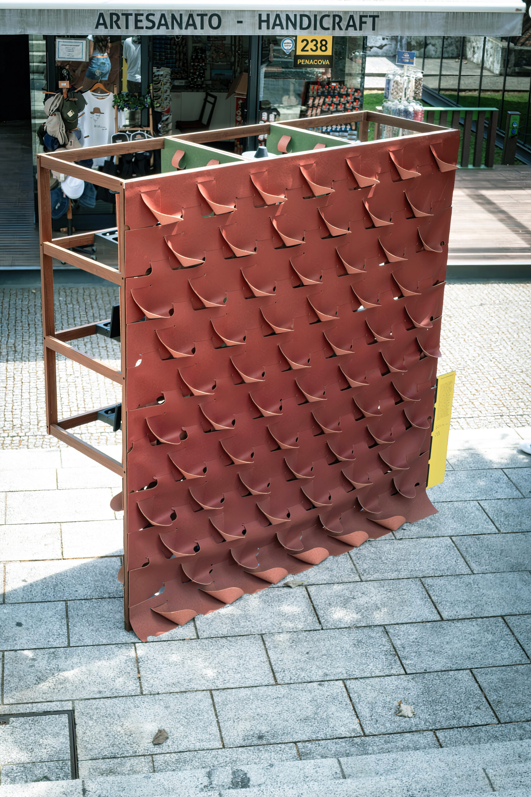 A think sheet of cork cut and folded in a wave-like design is positioned outdoors on a patio of light-colored stones. A shop in the background has a sign reading Artesanato - Handicraft.