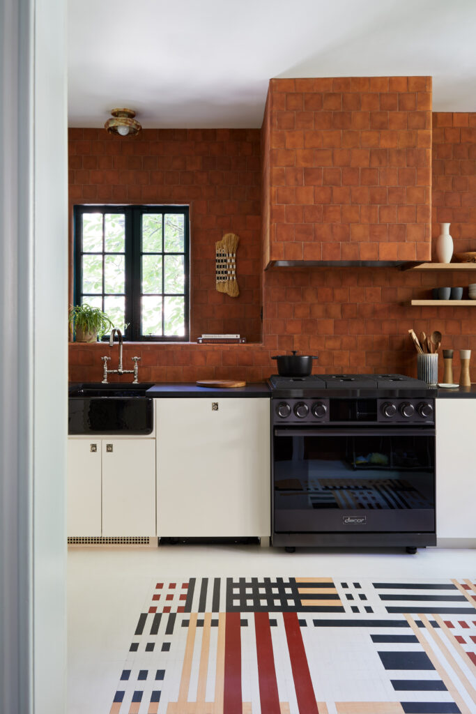 A kitchen with terra-cotta tiles decorating the wall, black and white cabinets, a black stove, and a floor painted with a blue, taupe, red, and white geometric pattern that resembles a woven textile.