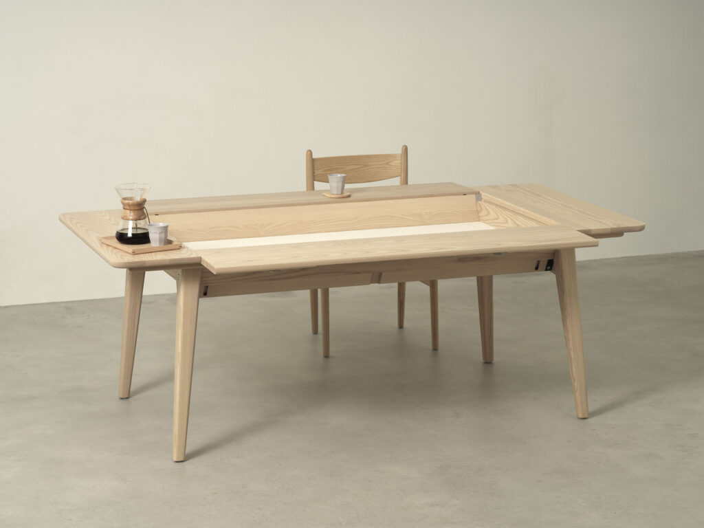 A rectangular pale wooden table with a single matching chair. The table features eaves around all sides and has a large depression in the center of the table. On the left eve is a serving tray with a single gray cup and a Chemex coffee maker filled with black coffee. In front of the chair is a coaster and a matching gray cup. 