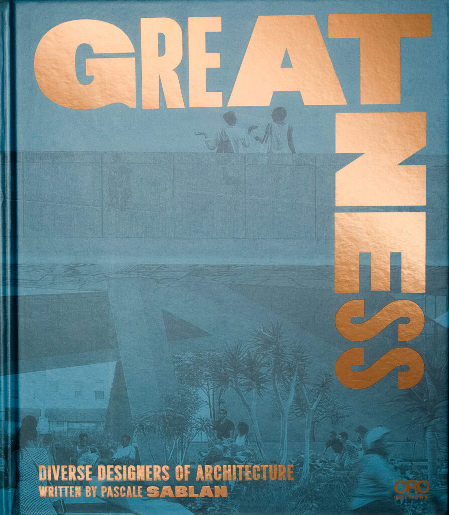 Book cover. Gold foil text with a bold, stylish typographical treatment reads “Greatness: Diverse Designers of Architecture Written by Pascale Sablan, Pro Editions.” The cover image, filtered in teal, depicts a modernist concrete walkway where two women engage in discussion. Below the walkway is a plaza where a mother sits with a young child, children run and play, and others stand. A speeding bicyclist can be seen in the bottom left corner of the photograph. 