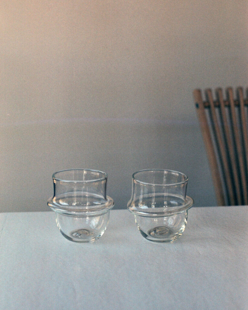 Two clear glasses rest on a table. The glasses have a conventional cylindrical top, but have glass rings around their centers. Below the glass rings, the glasses have a bubble-like form that tapers into a flat base.