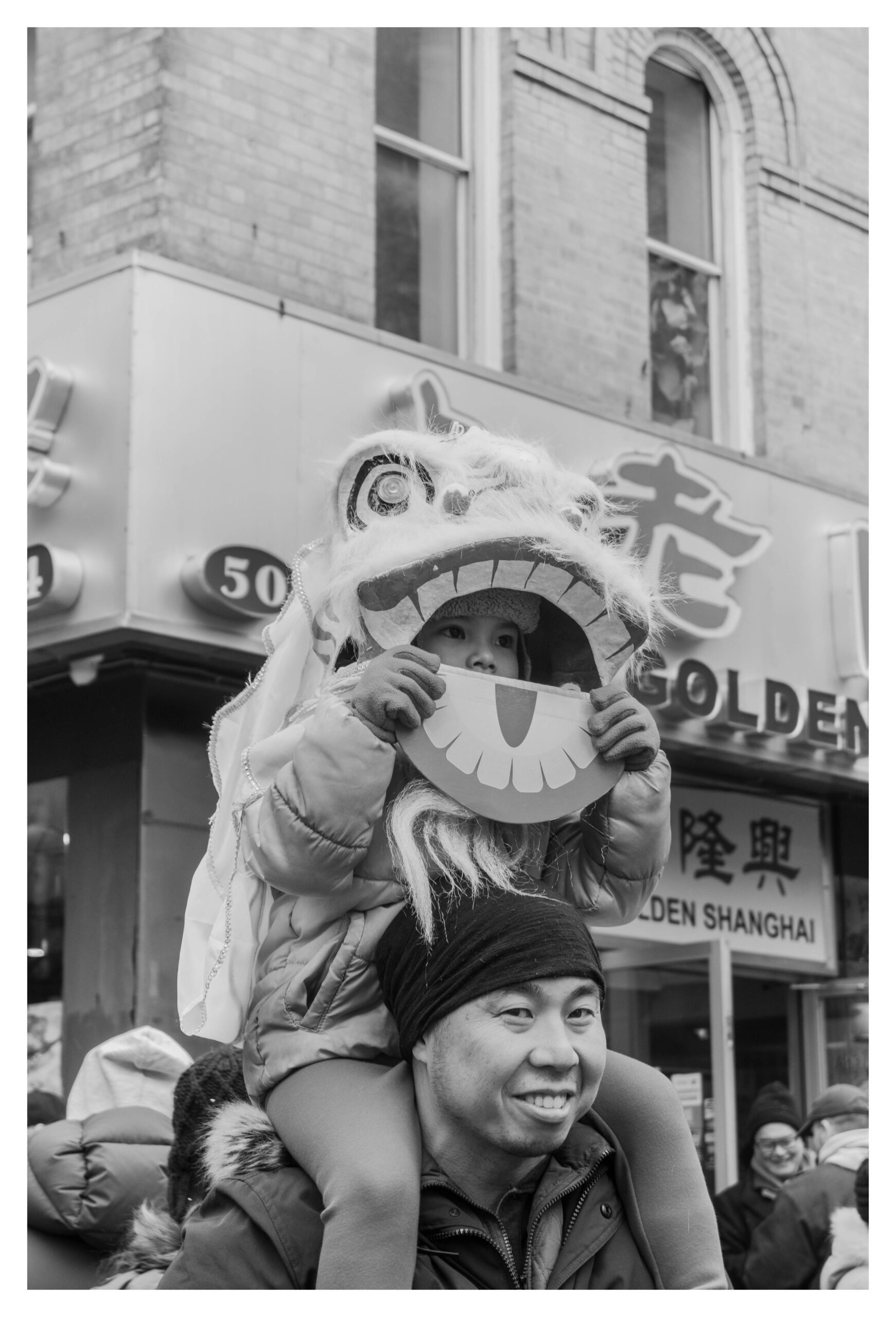 A black-and-white street scene of a man in a winter coat smiling as he carries a small child on his shoulders during a festival in Chinatown. The child, bundled in a puffy jacket, hat, and gloves, wears a furry lion dance costume head and holds a paper cutout shaped like a wide, toothy grin in front of their mouth. Behind them are storefront signs with Chinese characters and the words “GOLDEN SHANGHAI,” along with brick building windows and bundled onlookers.