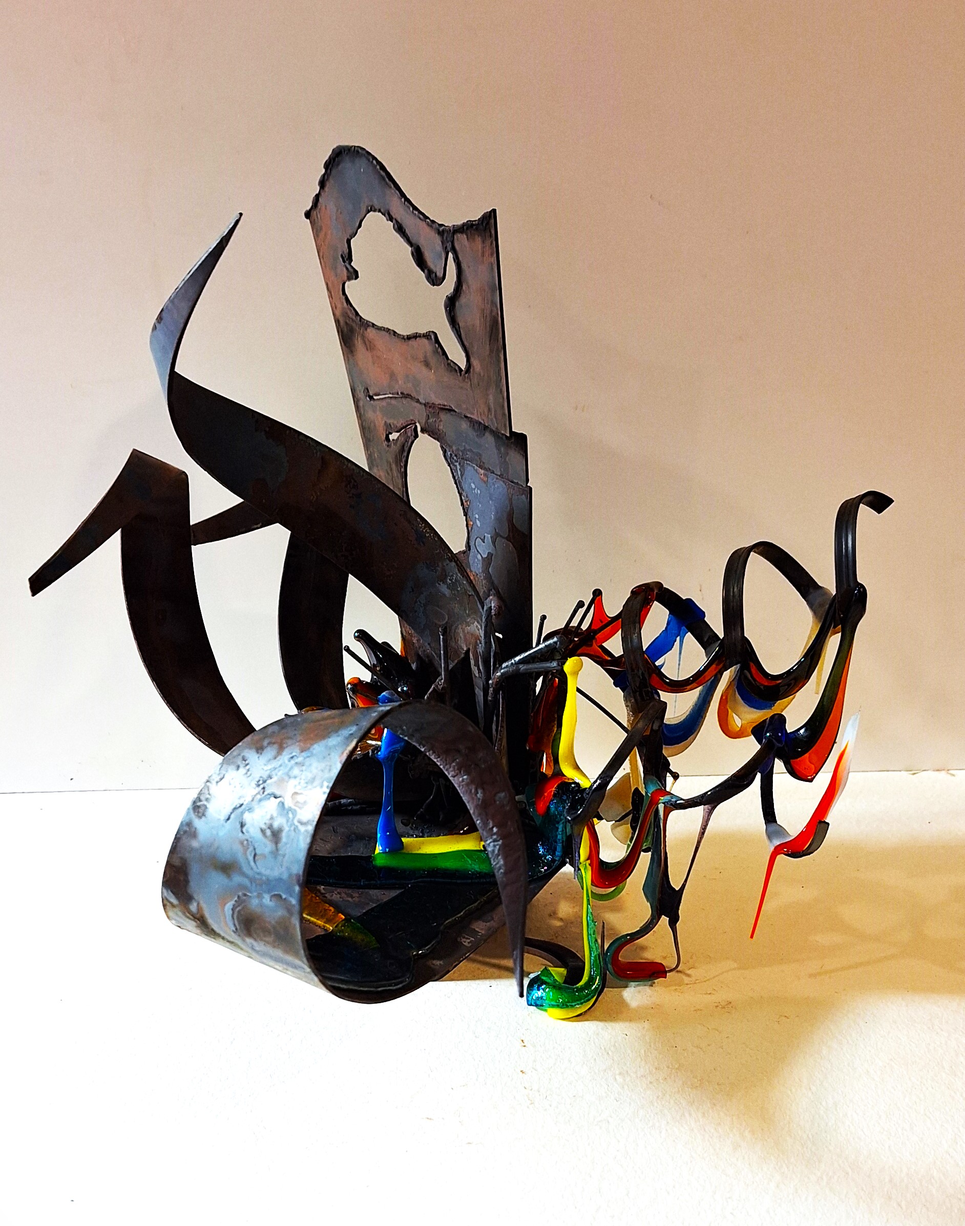 Small welded metal sculpture combining curved, twisted steel forms with brightly colored glass drips in red, blue, yellow, and green. The metal pieces arch, coil, and intersect, creating an industrial, almost calligraphic structure. Translucent glass accents cling to the steel like molten threads. The piece rests on a neutral tabletop against a plain beige wall, highlighting its rough textures and vivid color contrasts.