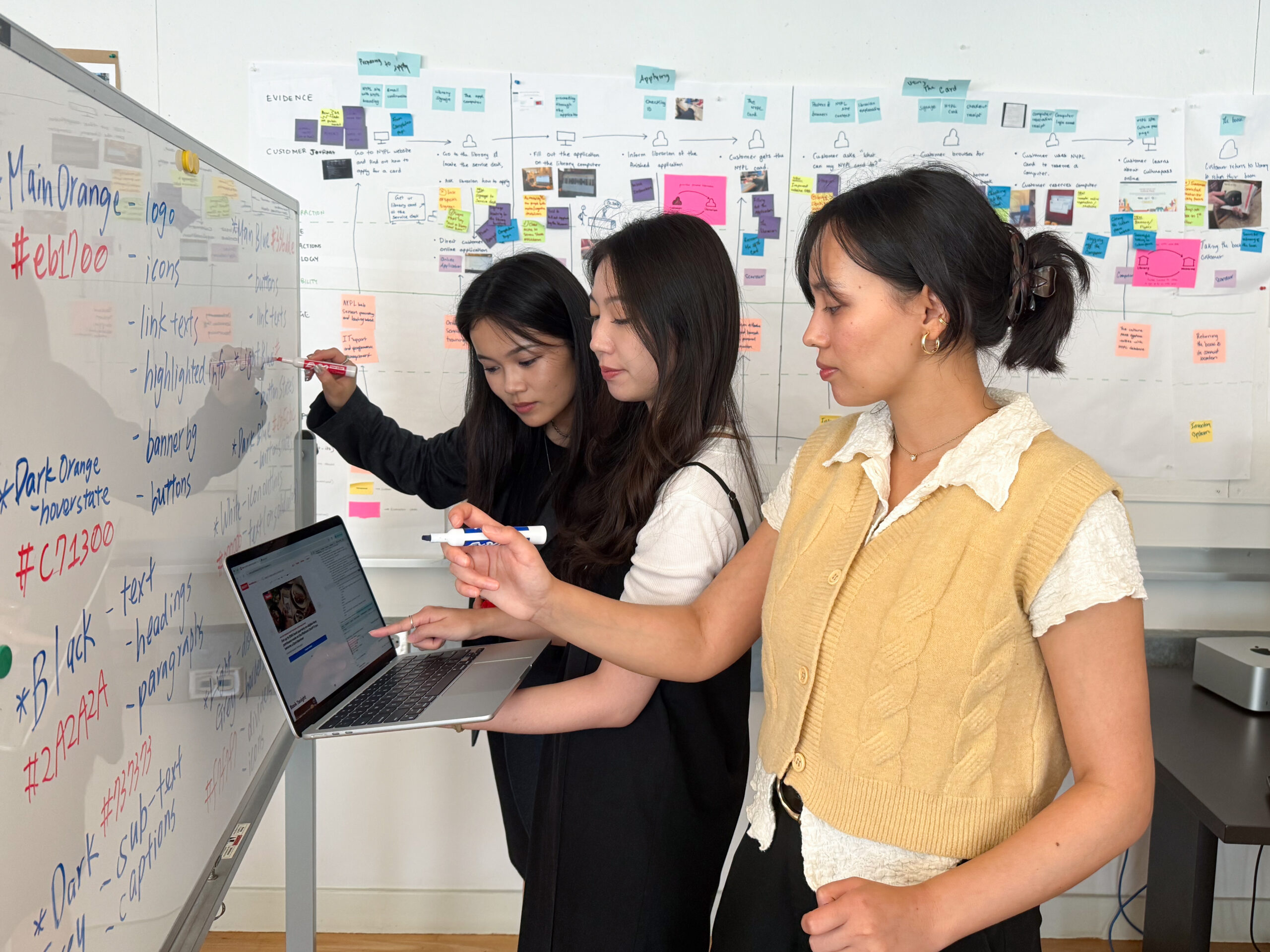 Three young women collaborate in a workspace. One writes on a whiteboard, another points at a laptop displaying a webpage, while the third stands beside them and holds a marker. Behind them, a wall is covered with colorful sticky notes and diagrams, suggesting a brainstorming session or project meeting. The environment appears informal and creative.