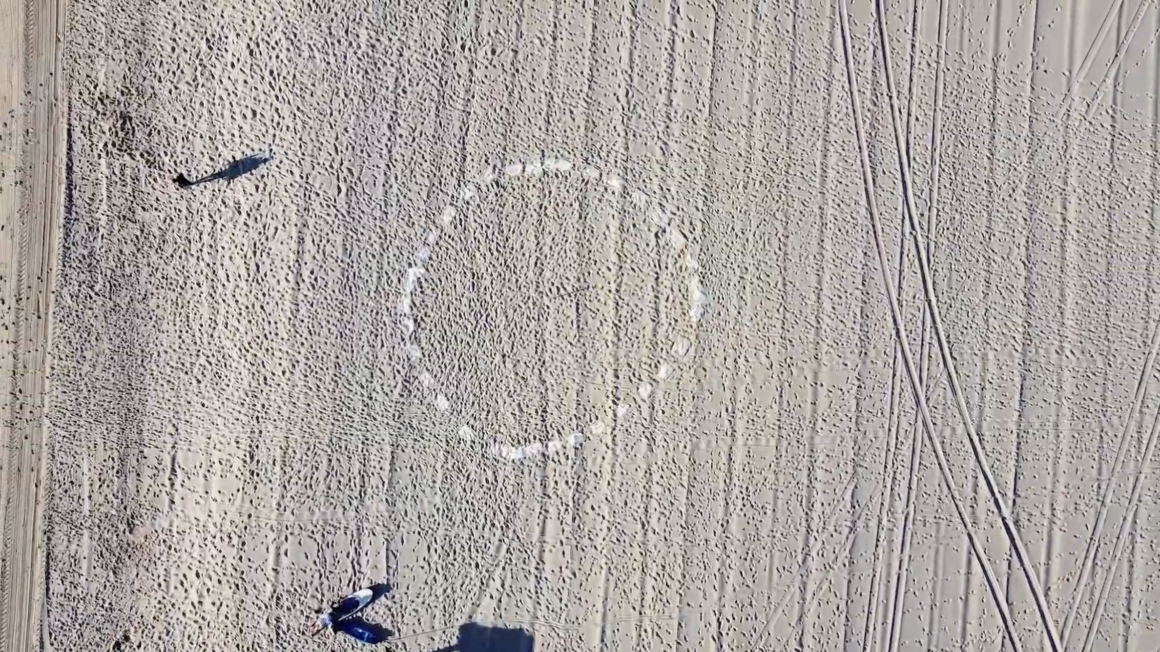 An aerial photograph of a sandy beach with visible footprints and textured ripples in the sand. At the center is a faint circular pattern made from small white markers arranged evenly in a ring. A long shadow from an unseen object extends across the left side of the frame. Two people stand near the lower left corner, looking toward the circle.