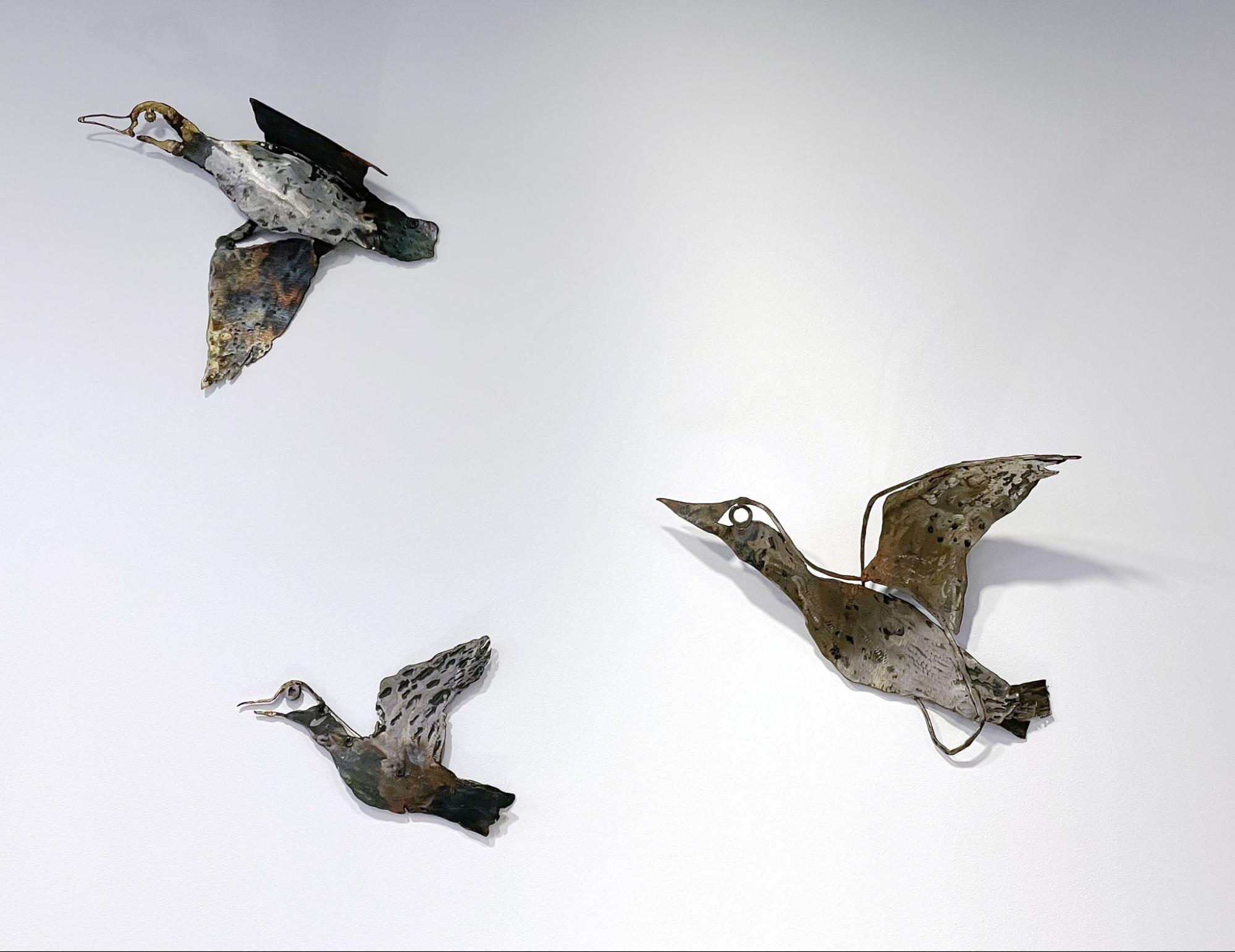 Three metal bird sculptures are mounted on a light wall. Each bird has different positions: the top bird is flying upwards with wings spread, the middle bird is ascending with wings slightly lifted, and the bottom bird is in a more horizontal position, appearing to glide. The sculptures feature a mix of browns, greens, and metallic tones, adding a rustic charm.