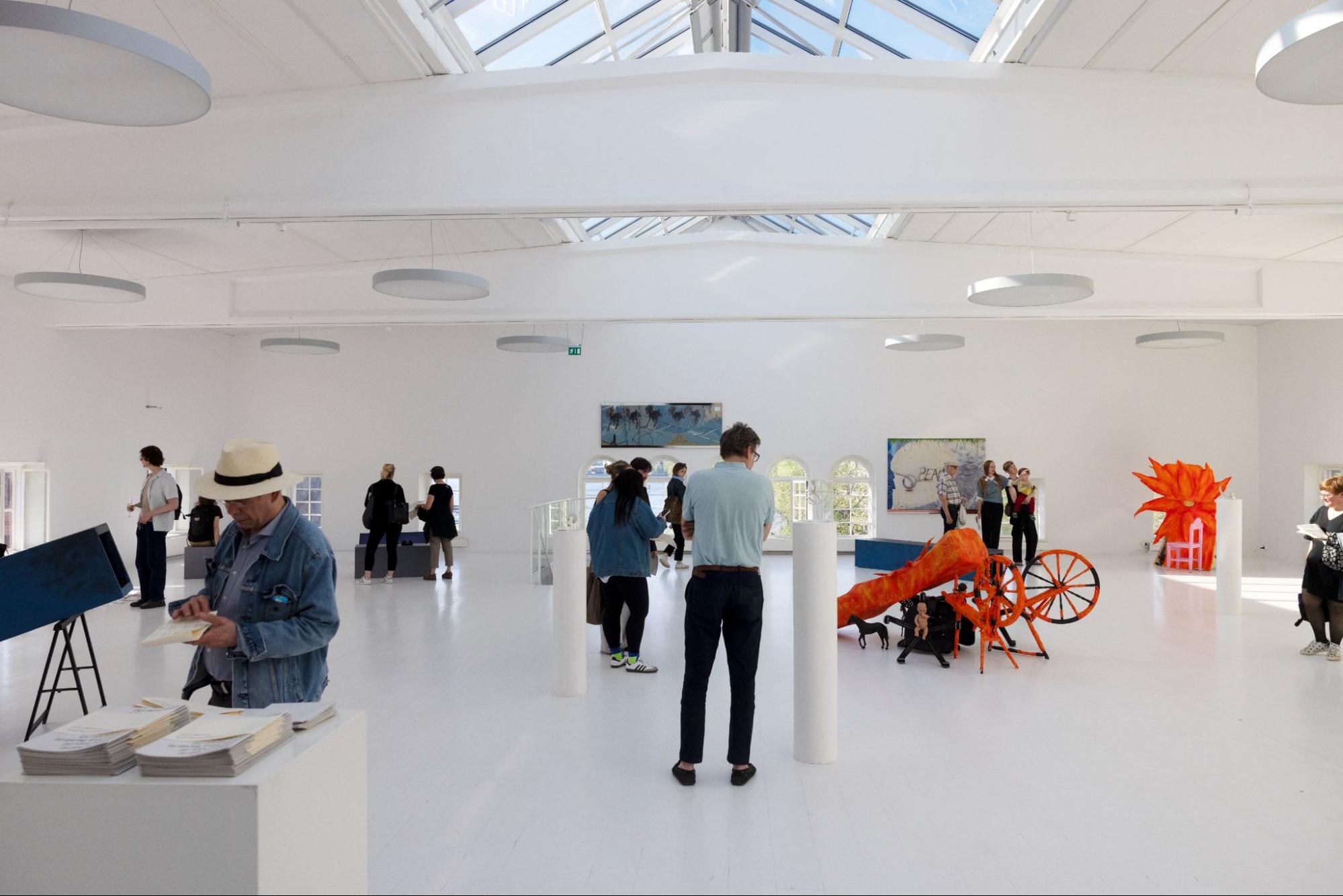 A bright white gallery interior with a large skylight illuminating visitors who observe artwork, sculptures, and installations spaced throughout the open room.