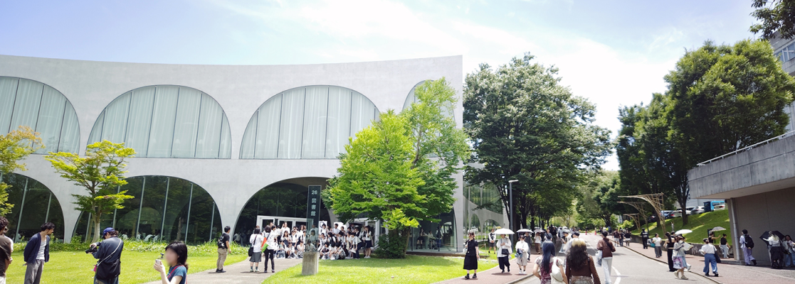A wide exterior view of a modern building with large curved window openings and smooth white concrete walls. People walk and gather on a sunny day, standing on grass and paved walkways surrounded by tall green trees.
