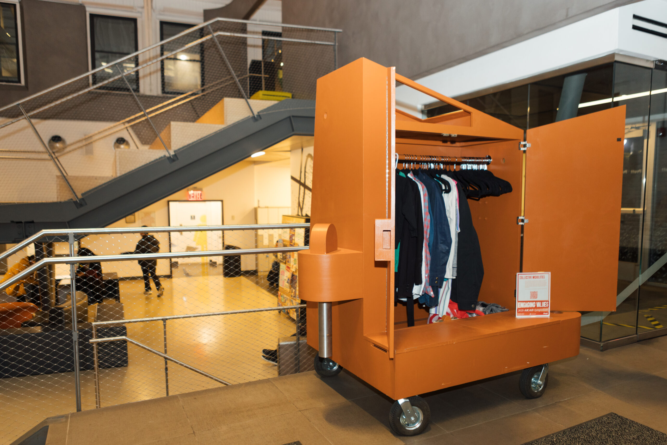 An orange, wheeled clothing rack is positioned near a staircase in a modern indoor space. The rack has an open door displaying various shirts and jackets hanging on hangers. In the background, people are seen engaging in conversation, and there's a door leading to another room. The overall environment appears to be a community or social area with a contemporary design.