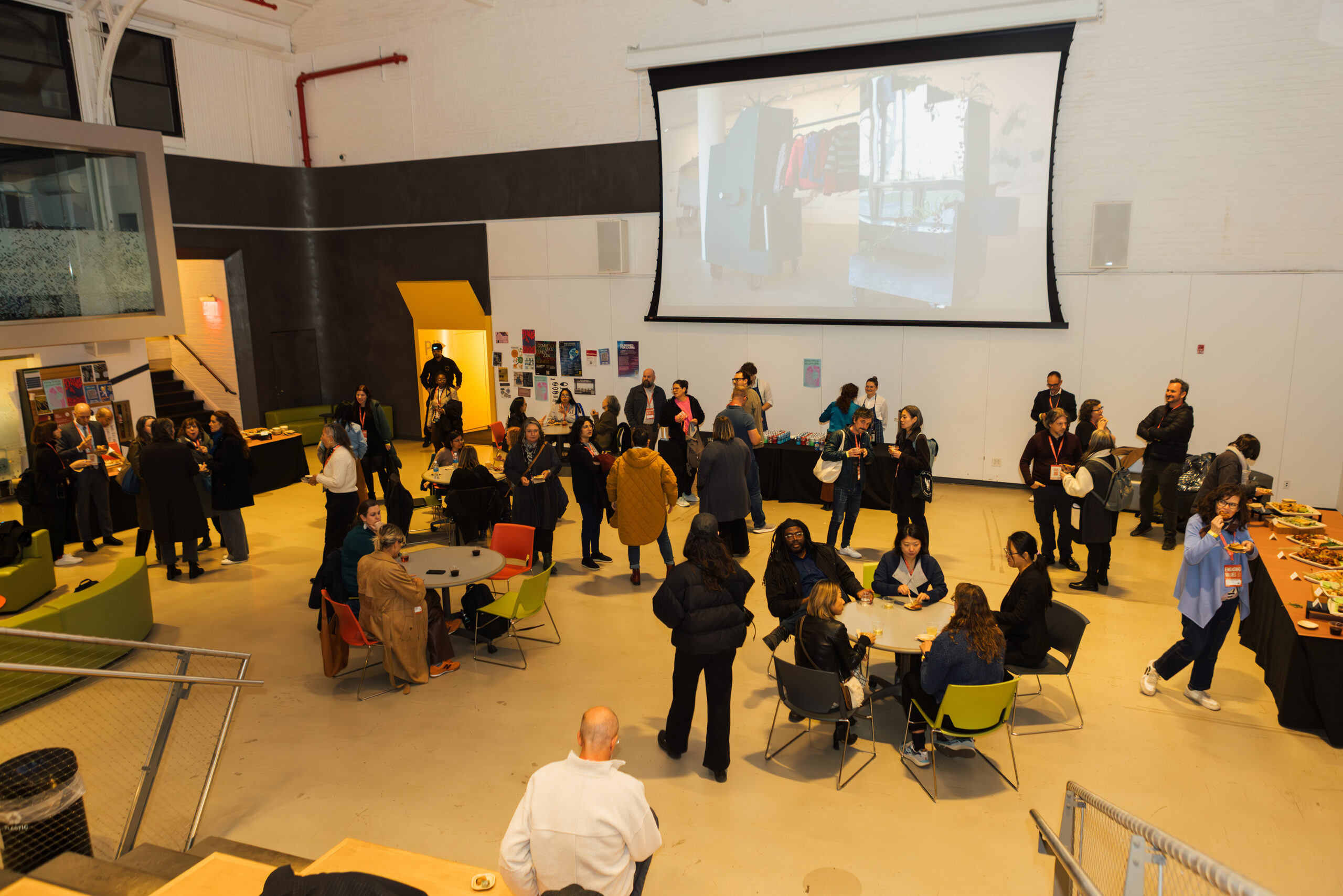 A spacious indoor venue filled with people attending an event. Groups of attendees socialize and enjoy refreshments at tables and standing. Food is displayed on a long table, and a presentation is visible on a large screen in the background. The décor features colorful seating and posters on the walls.