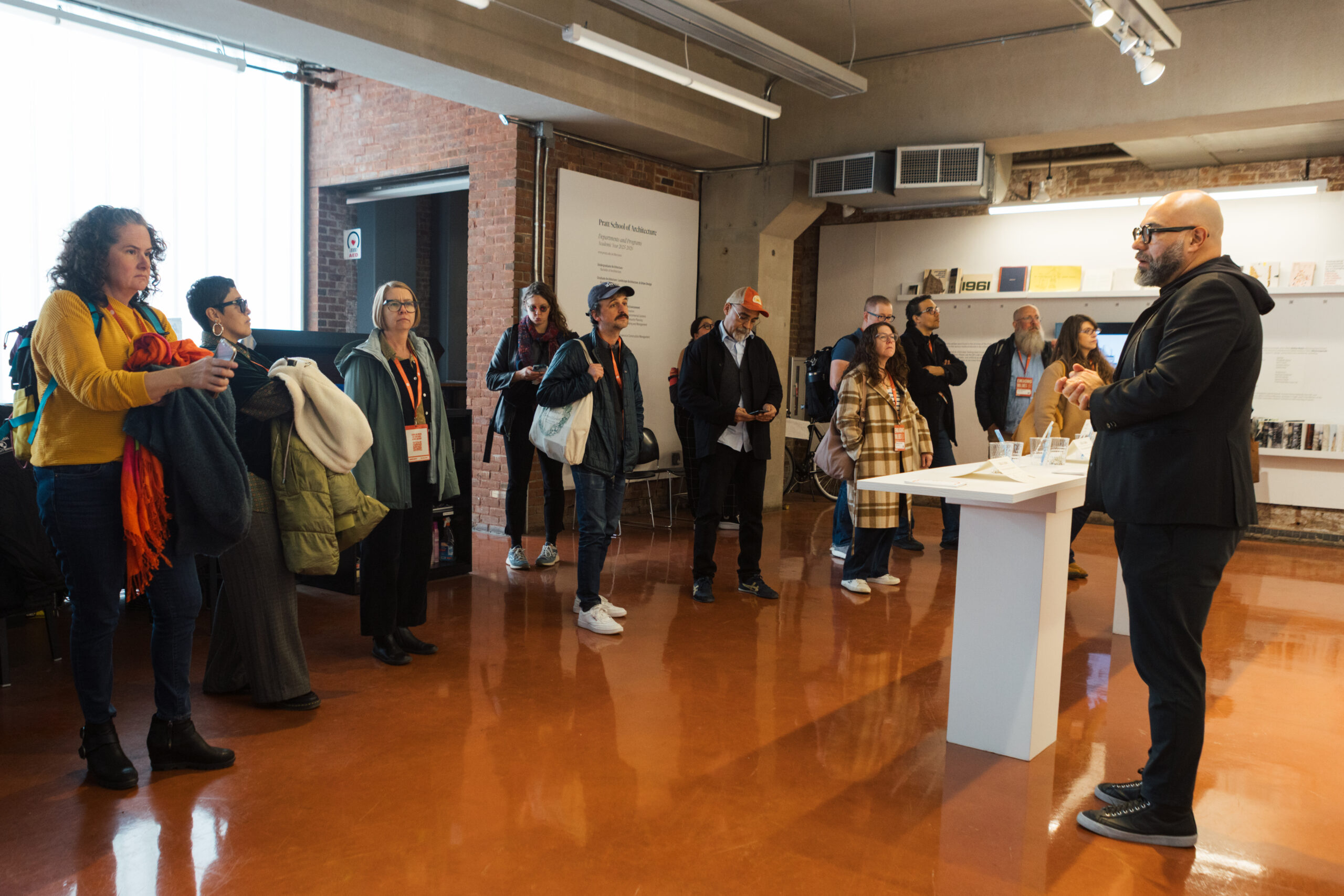 A group of people is gathered in a well-lit room with brick walls, listening attentively to a speaker. The speaker, a man in a black suit, stands at a table displaying materials. The audience, consisting of diverse individuals, includes women and men, some holding their belongings and others engaged with their phones. There are books and materials on display in the background, enhancing the educational atmosphere.