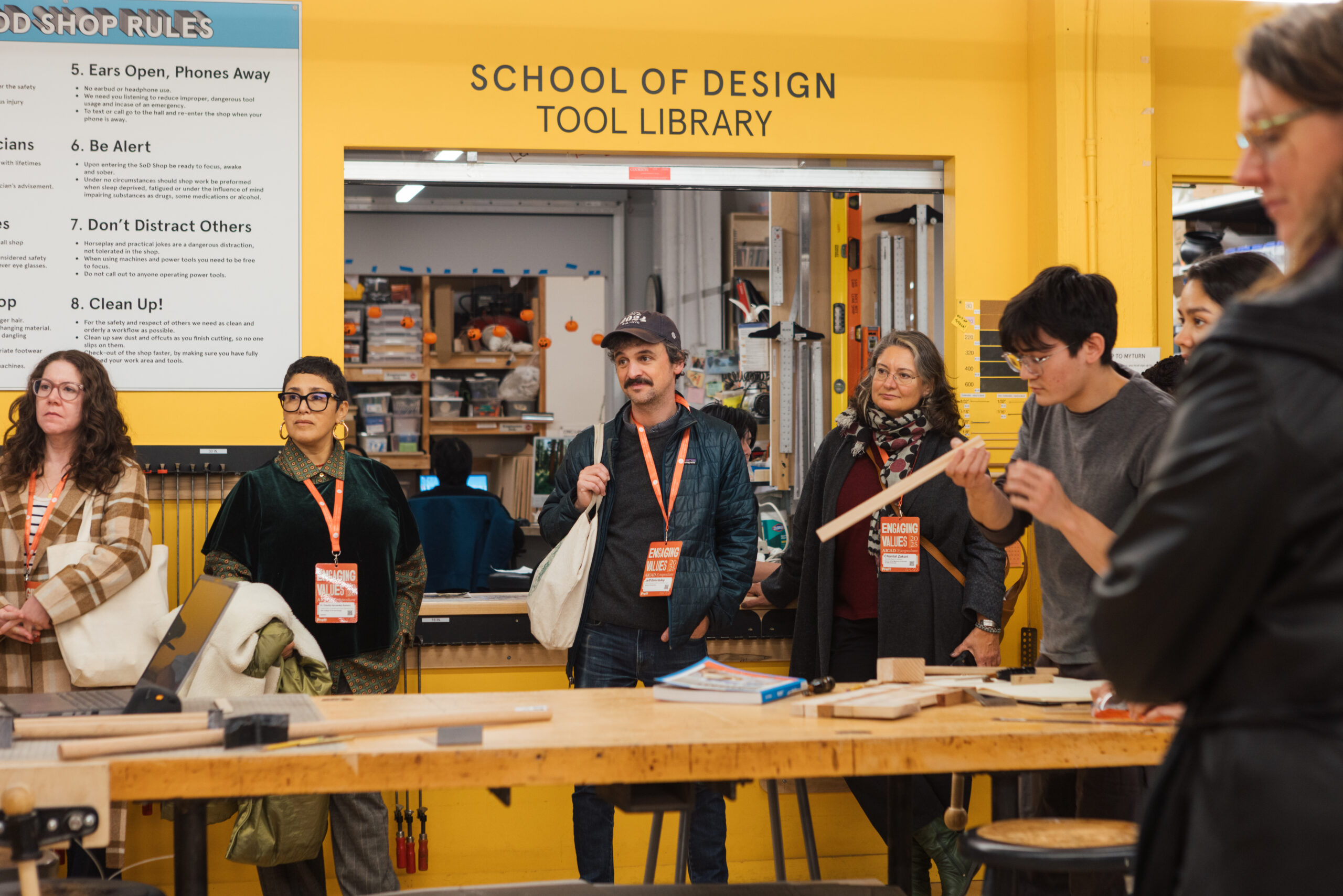 A group of people stands in a brightly colored workshop, engaging in discussion. The background features a sign listing woodshop rules on the left, while the entrance to a tool library is above. The individuals wear casual attire and name tags, with two people focused on a piece of wood. The atmosphere suggests a creative or educational setting related to design or woodworking.