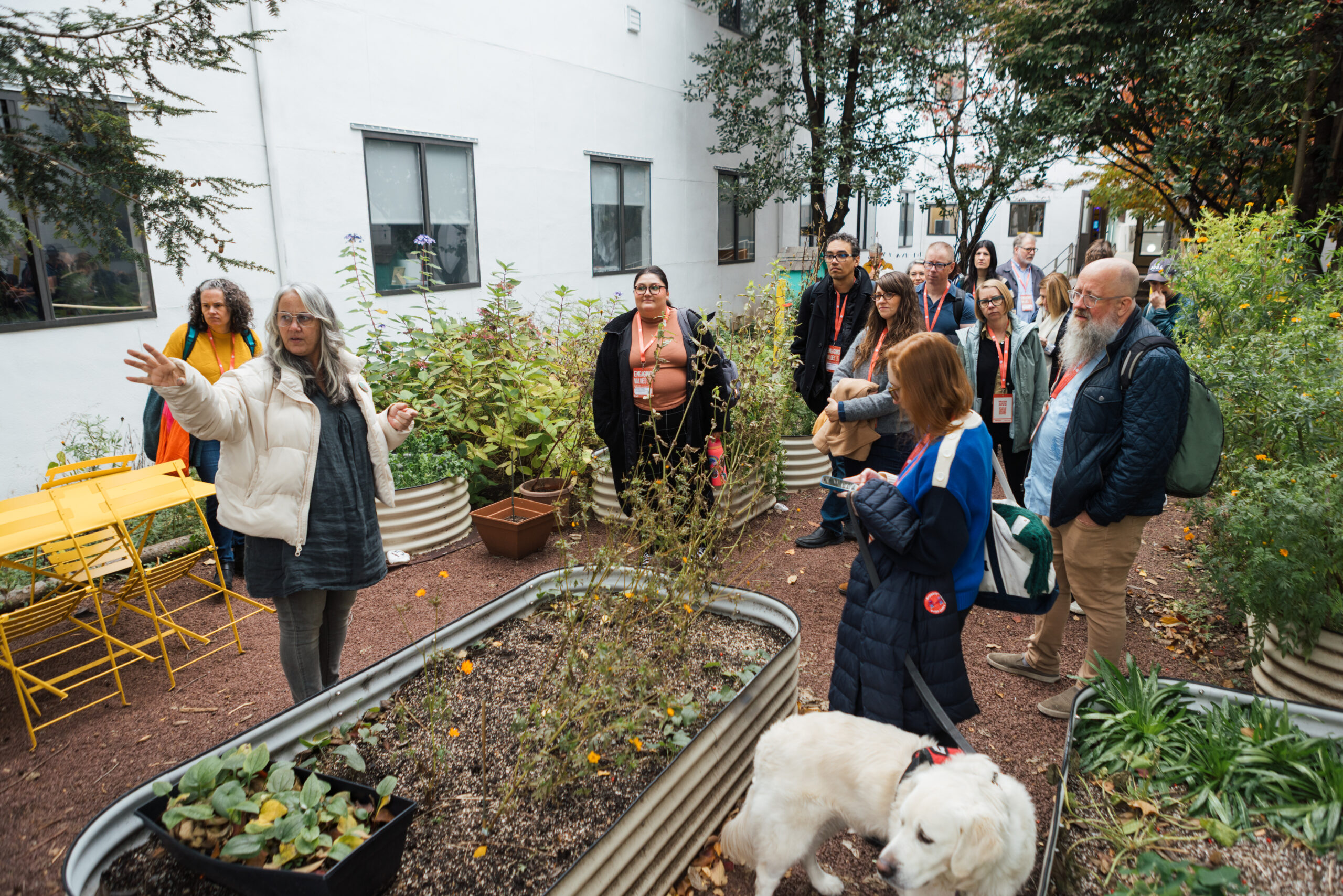 A group of individuals gather in a brightly colored workshop space labeled "School of Design Tool Library." They appear engaged in a discussion. A list of shop rules hangs on the wall behind them. One male participant is holding a wooden piece, while others listen attentively. The atmosphere is collaborative and informative, with various tools visible in the background.