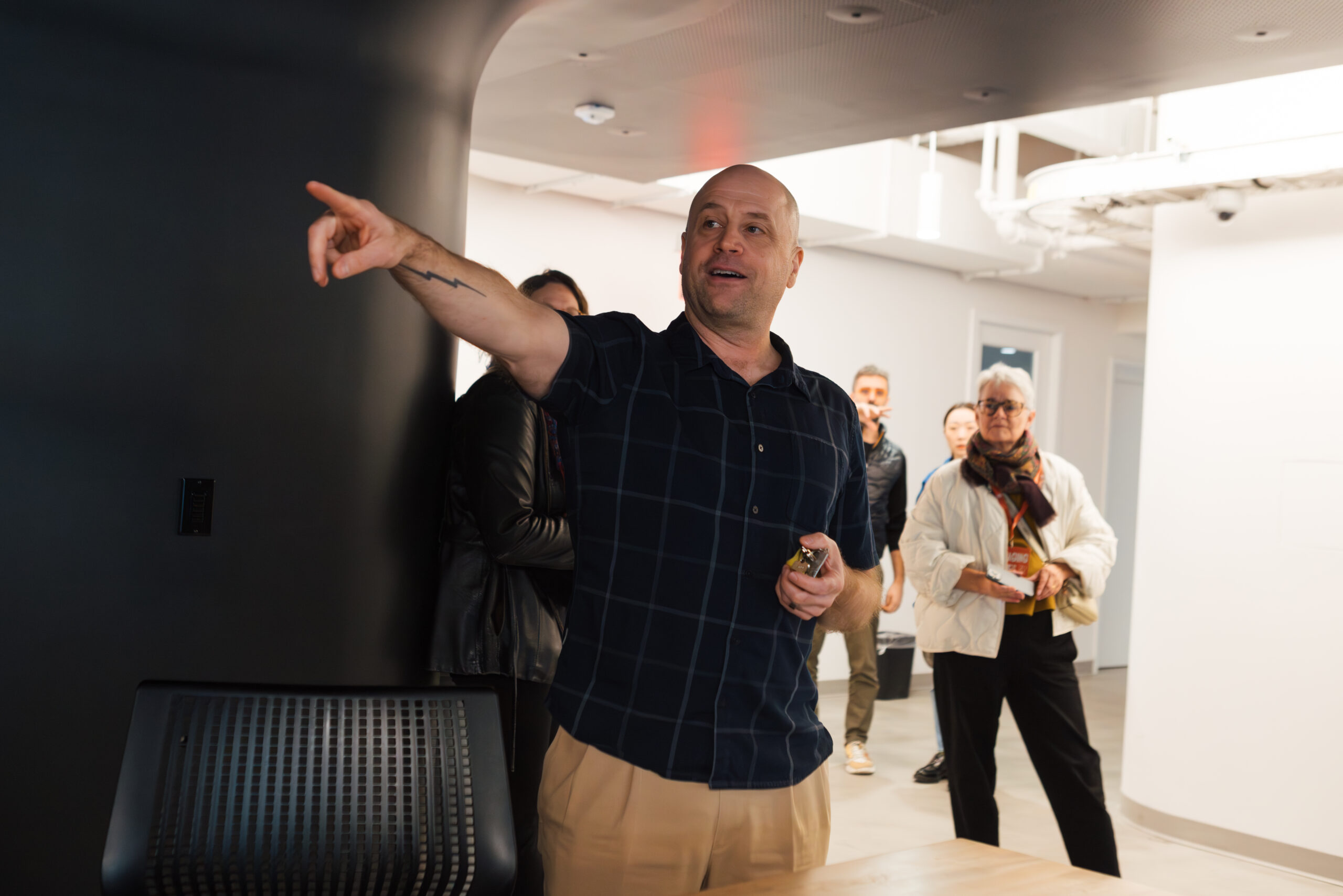 A man with short, bald hair and a tattoo on his arm gestures while speaking to a group. He wears a dark plaid shirt and beige pants. In the background, several people listen attentively, including a woman in a white jacket and glasses. The setting appears to be a modern office space with white walls and a black accent wall. A black chair is visible in the foreground.