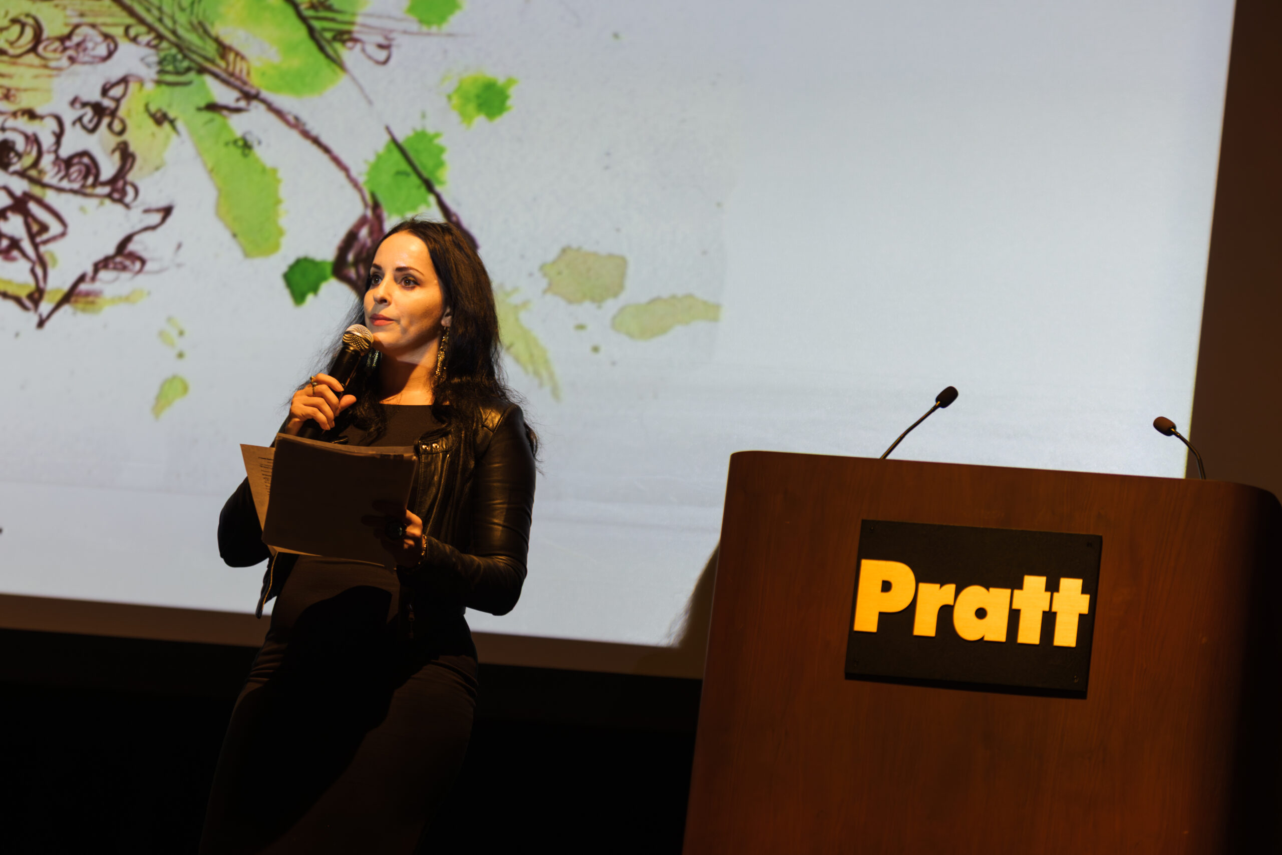A woman stands at a podium with the "Pratt" logo, holding a microphone and papers. She has long dark hair, wears a black leather jacket, and looks towards the audience. A colorful, artistic background featuring green and brown elements is projected behind her, suggesting a creative or artistic presentation.