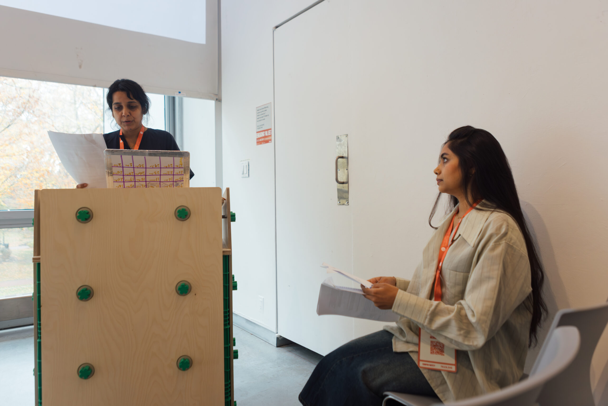 A woman stands behind a wooden podium reading from a document, with a notepad in front of her displaying data. Nearby, another woman sits on a chair, holding pages and looking attentively at the speaker. Both are in a modern, well-lit room with large windows and minimalistic decor.